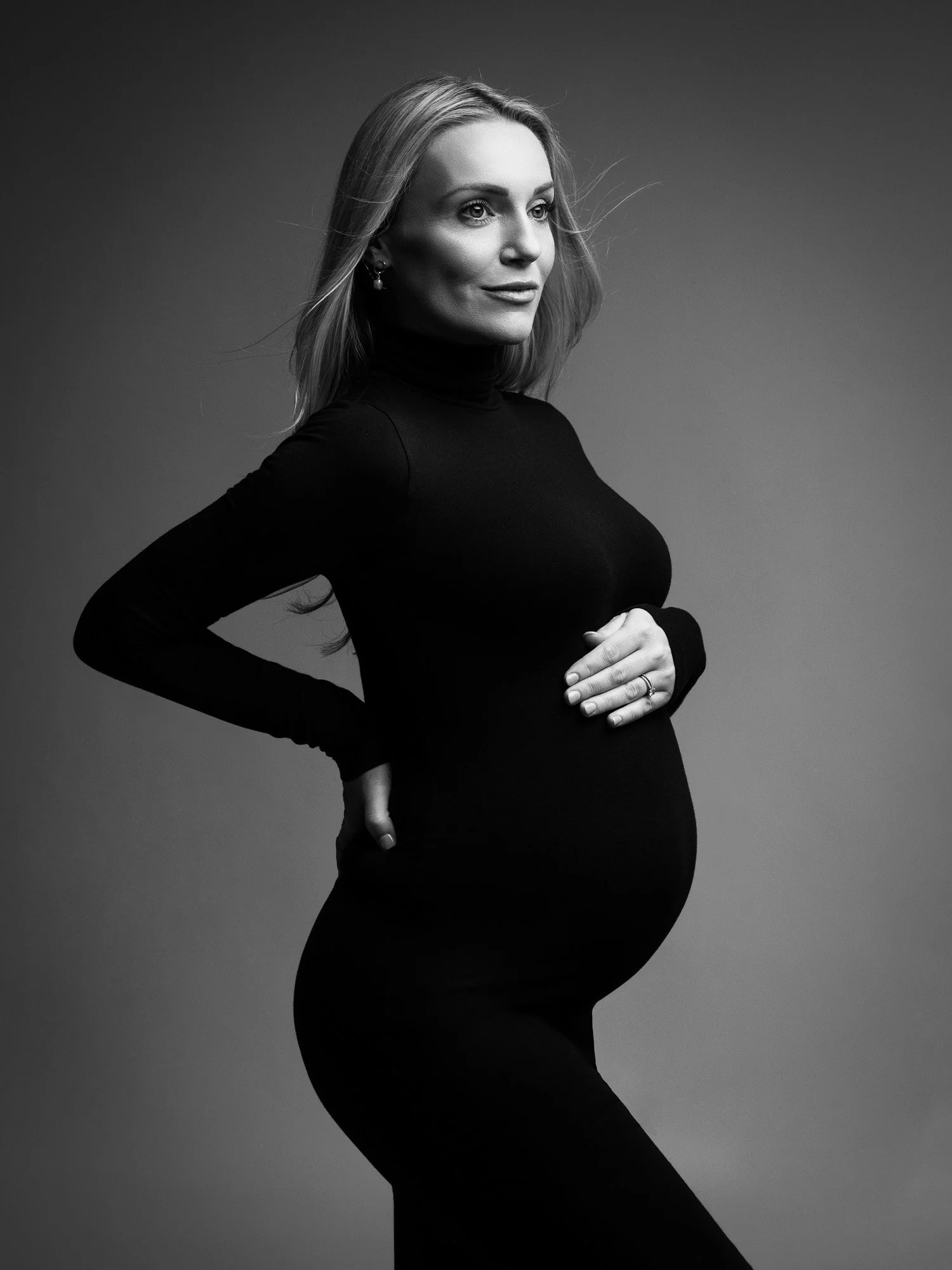 Black and white studio portrait of a pregnant woman in a black turtleneck, looking to the side with one hand on her belly and the other on her back.