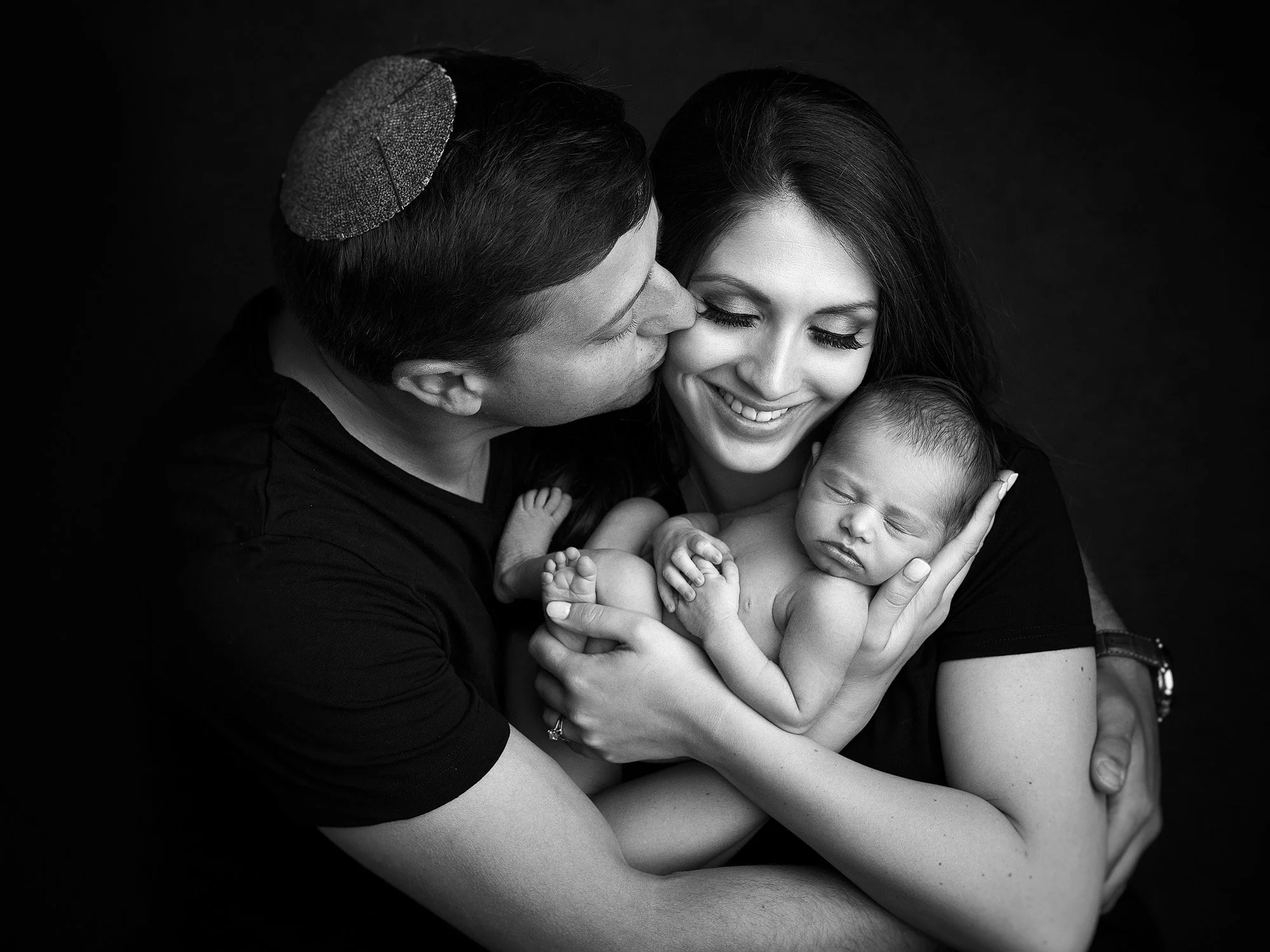 Black and white photo of a family with a father, mother, and newborn baby, with the father giving a gentle kiss on the mother's cheek while she holds the sleeping baby.