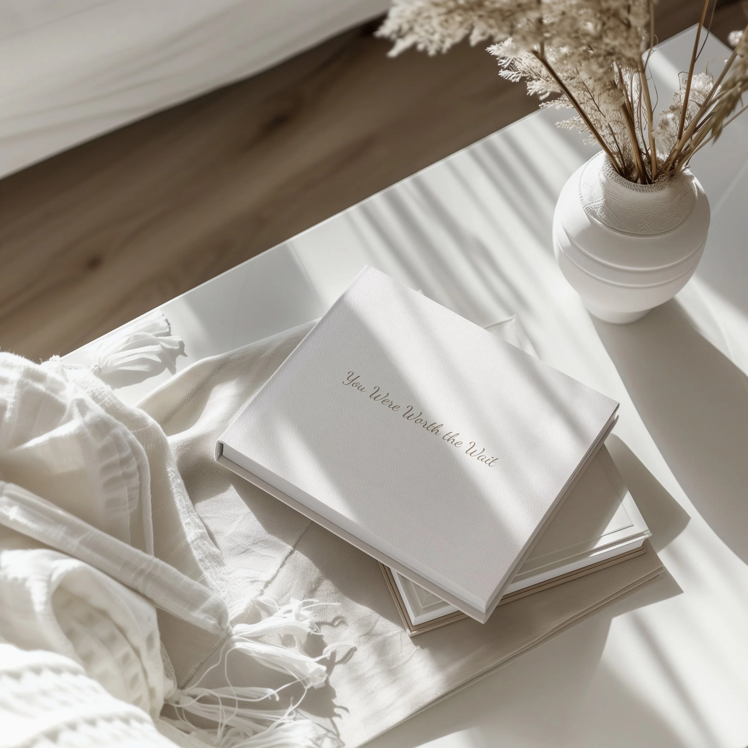 A white photo album with the words "You Were Warth the Wait" embossed on the cover, resting on a white cloth and a white surface. Next to the album is a vase with dried flowers, casting shadows in sunlight.