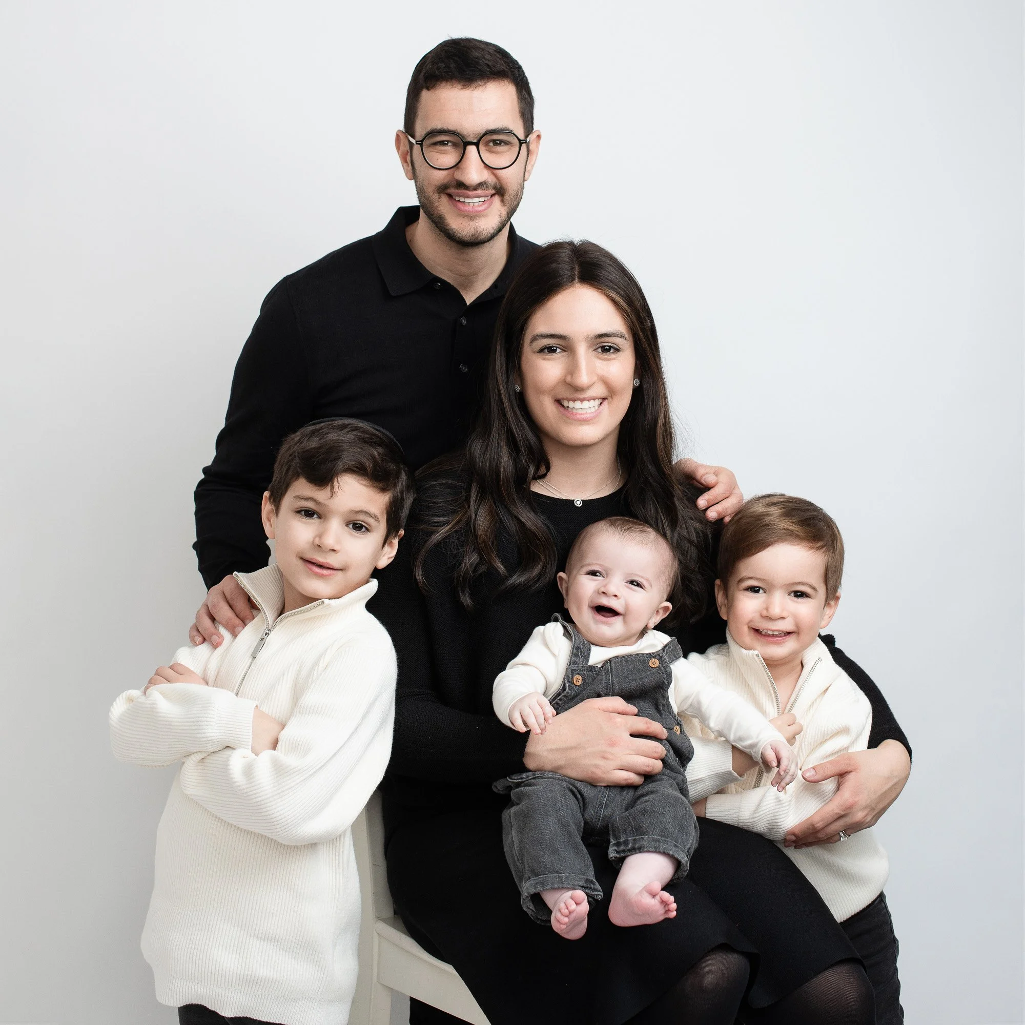 A family of six posing together against a plain white background. The father is standing at the back, wearing glasses and a black shirt. The mother is sitting in the center, holding a baby in her lap, and is wearing a black top. Two young boys are st