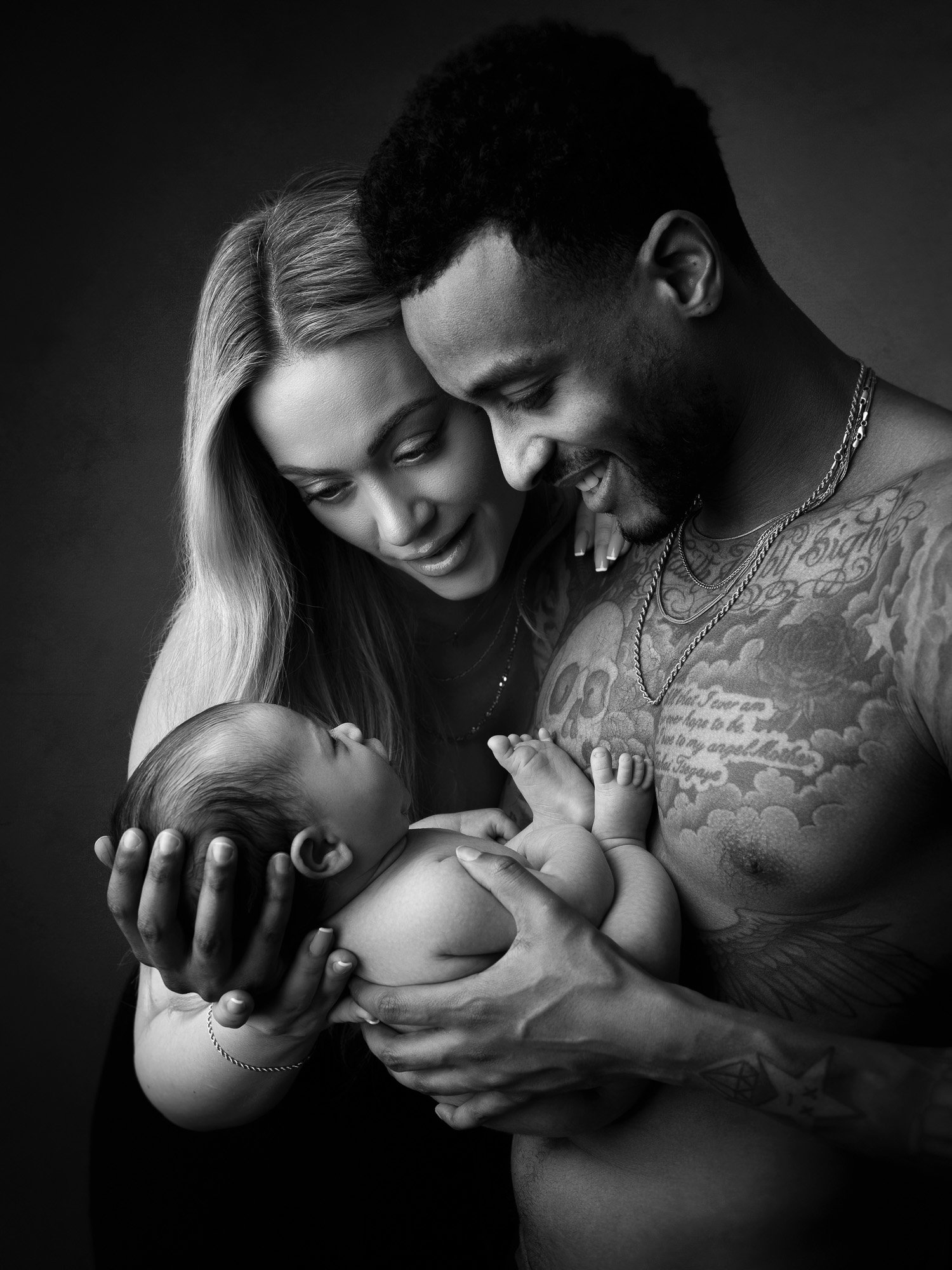 Black and white photo of a family: a woman and man, both smiling, holding their newborn baby close to their chests.