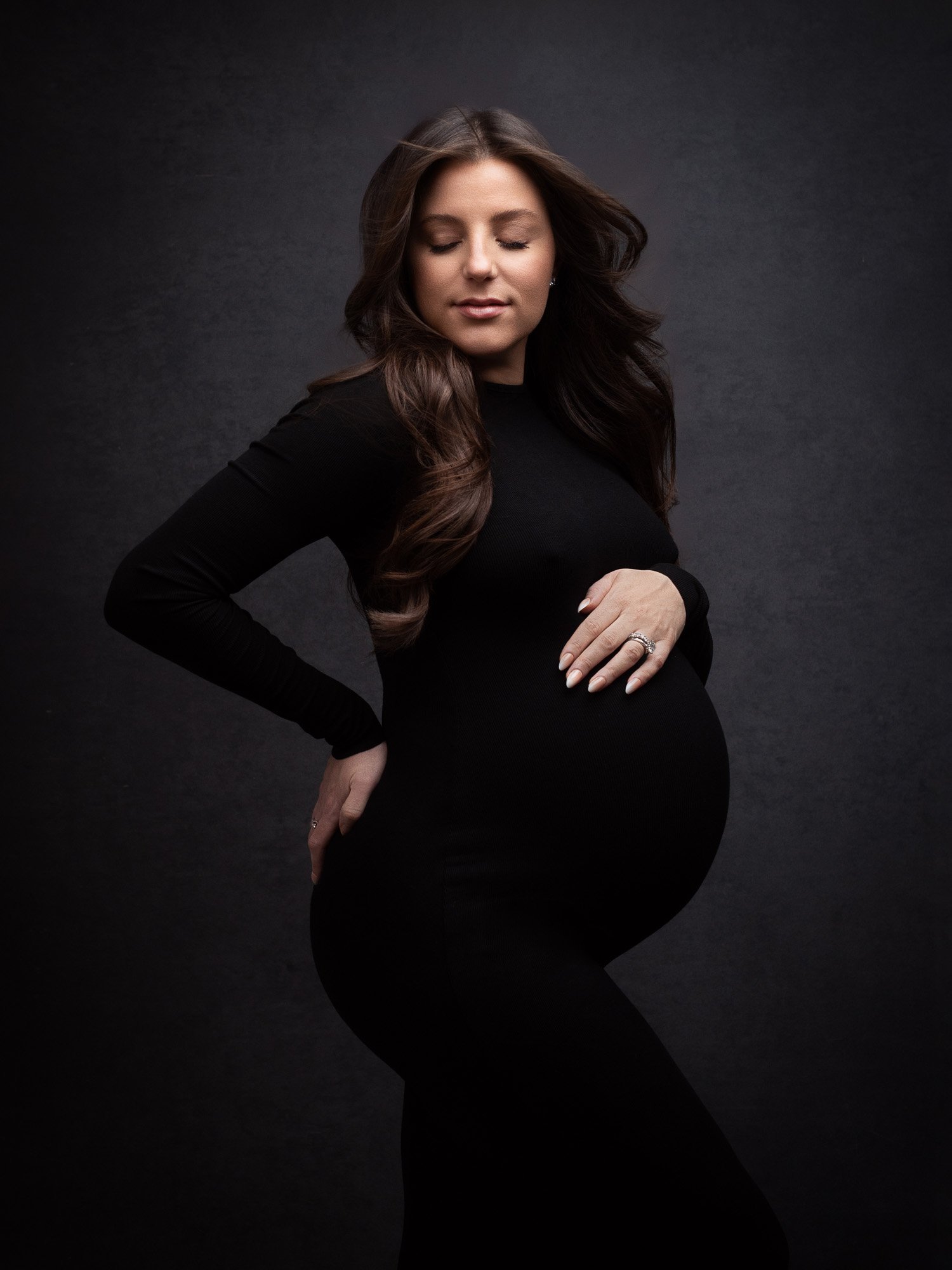 A pregnant woman in a black dress posing with her eyes closed against a dark background.