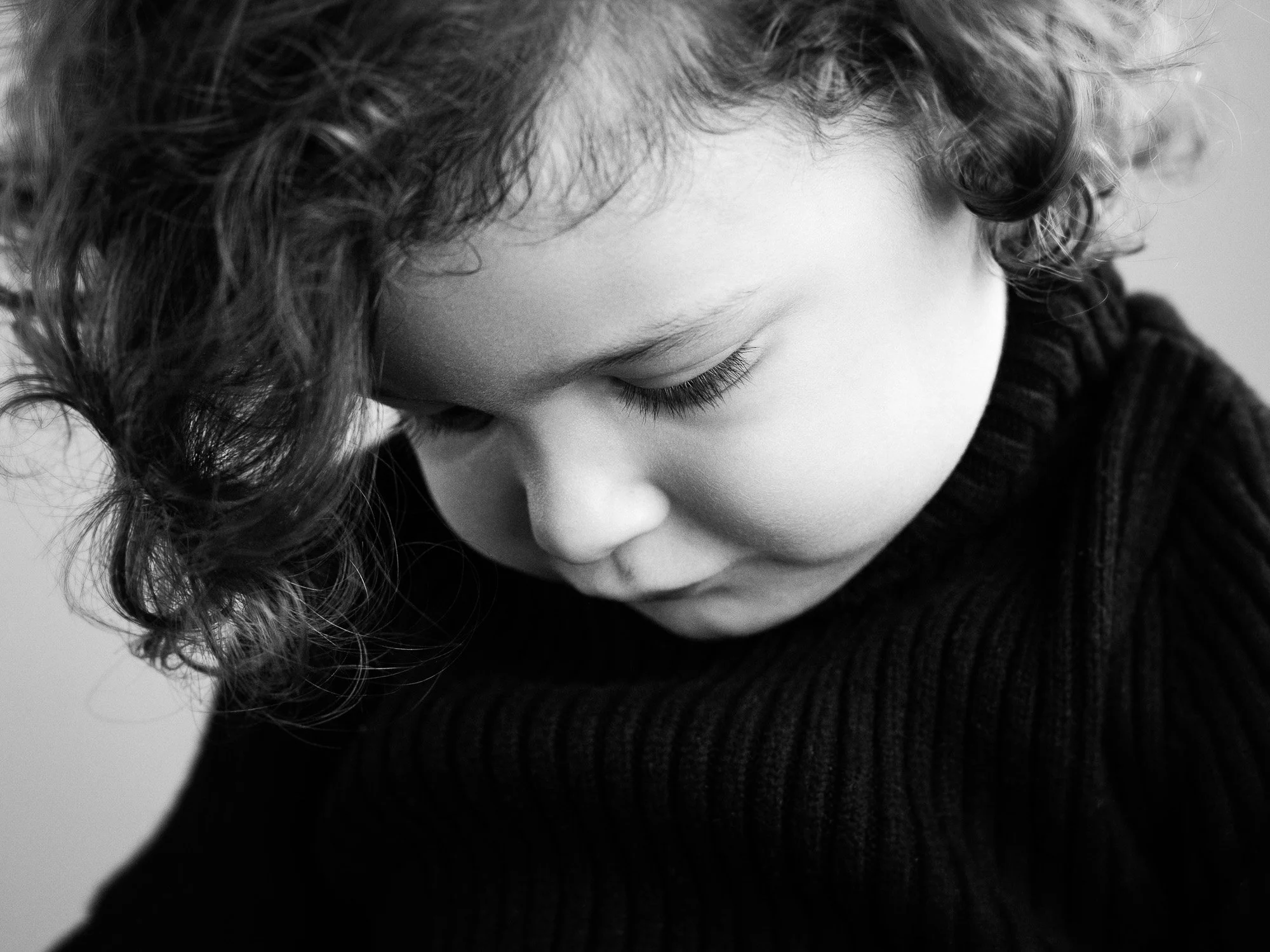 Close-up of a young child with curly hair and a black sweater, looking down with eyes closed.