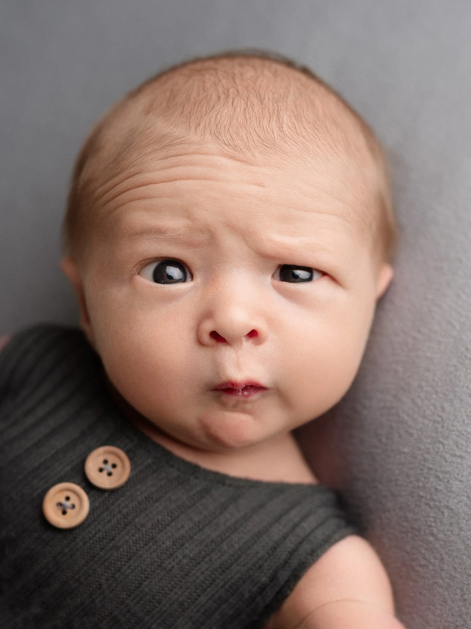 Close-up of a baby with big eyes and a curious expression, wearing a dark sleeveless shirt with two buttons, lying against a grey background.