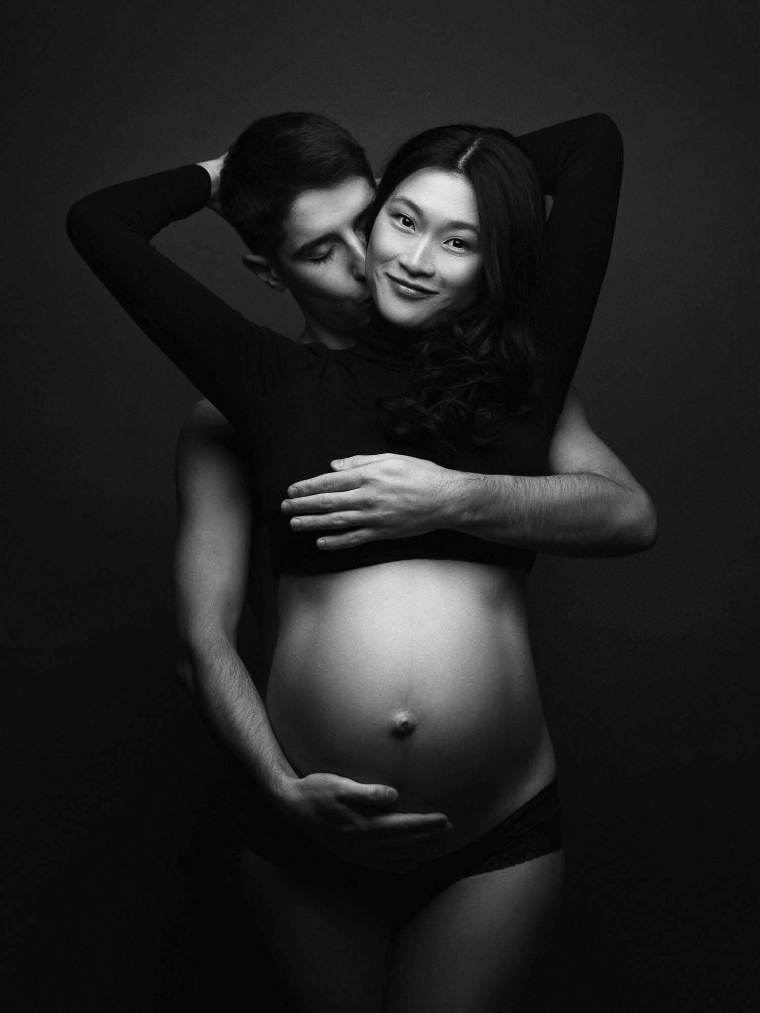 A black-and-white photo of a pregnant woman with long dark hair, smiling, wearing a black top and undergarments, with a man behind her, kissing her cheek and holding her belly. The man has short hair and light skin, and his face is partially hidden.