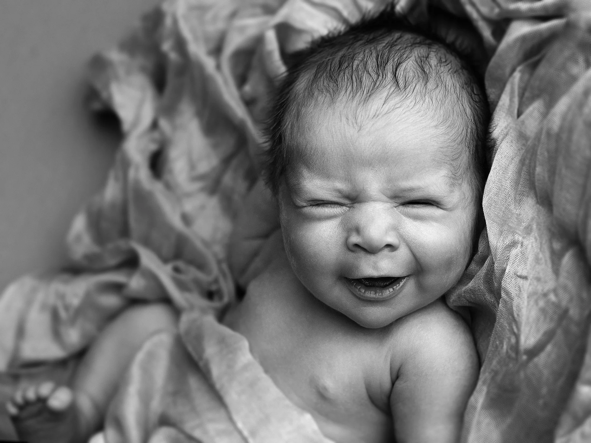A black and white photo of a baby with a big smile, lying on a textured blanket.