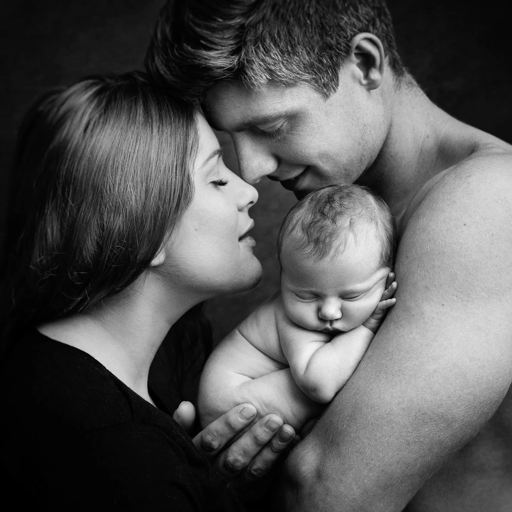 A black and white photograph of a couple holding their sleeping baby close with their foreheads touching, showcasing a tender moment of love and bonding.
