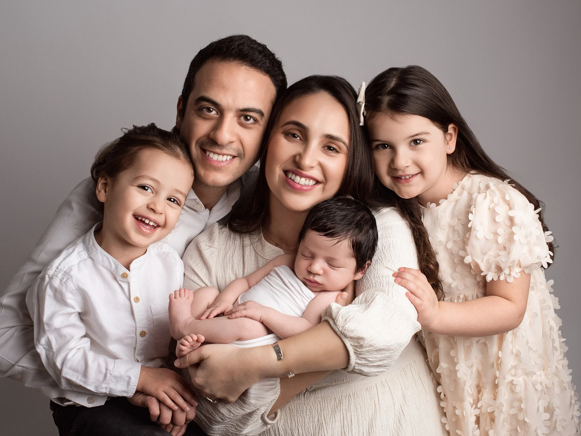A smiling family of six posing closely together in a studio, including two adults, two young girls, a young boy, and a newborn baby.