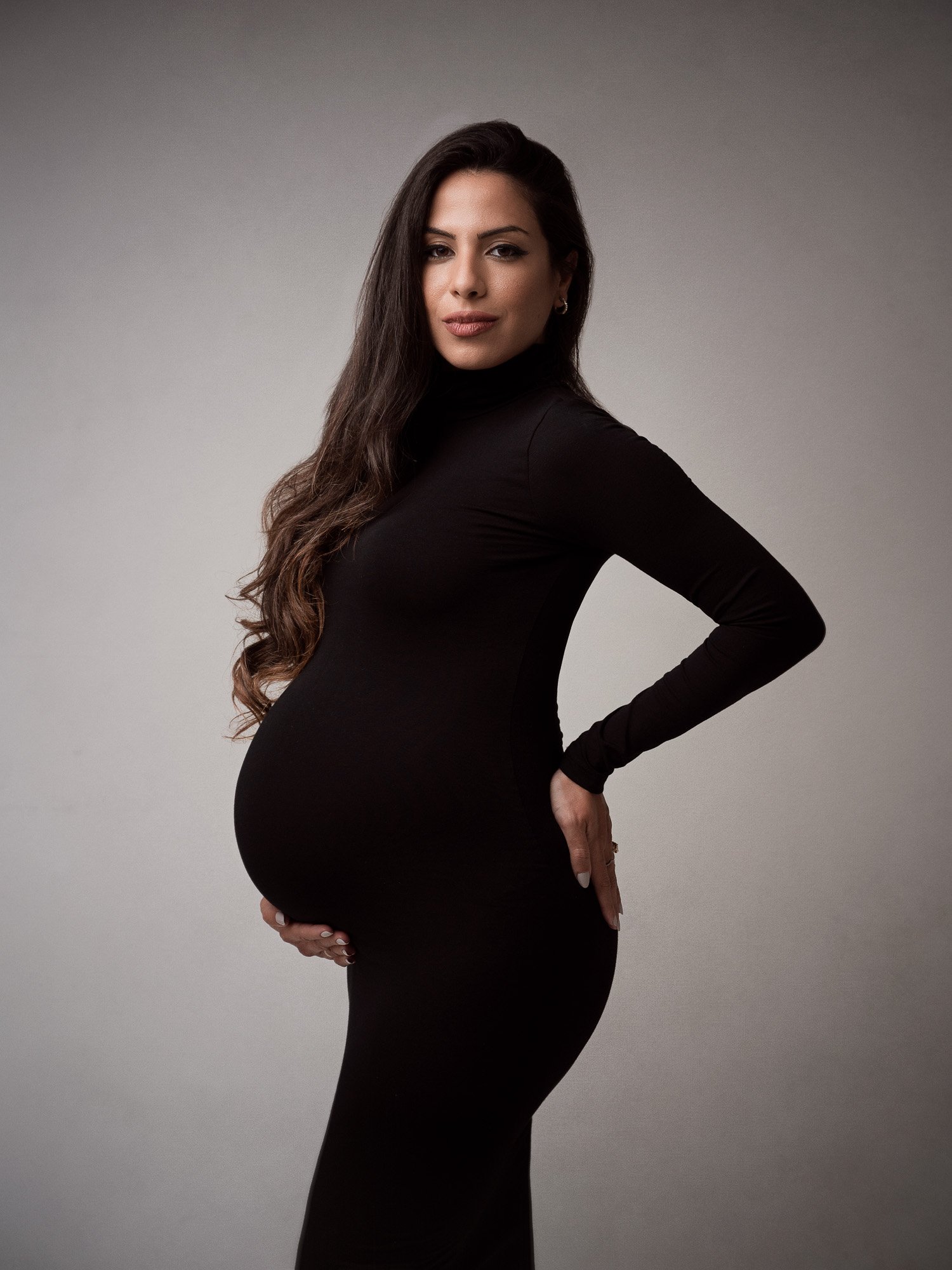 Pregnant woman with long dark hair in a fitted black dress posing against a plain background.