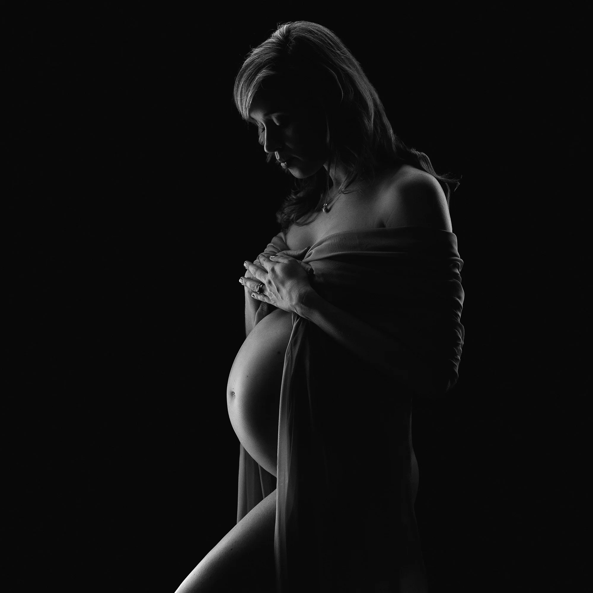 Silhouette of a pregnant woman with hair down, softly holding her belly with both hands, draped in a cloth, against a black background.