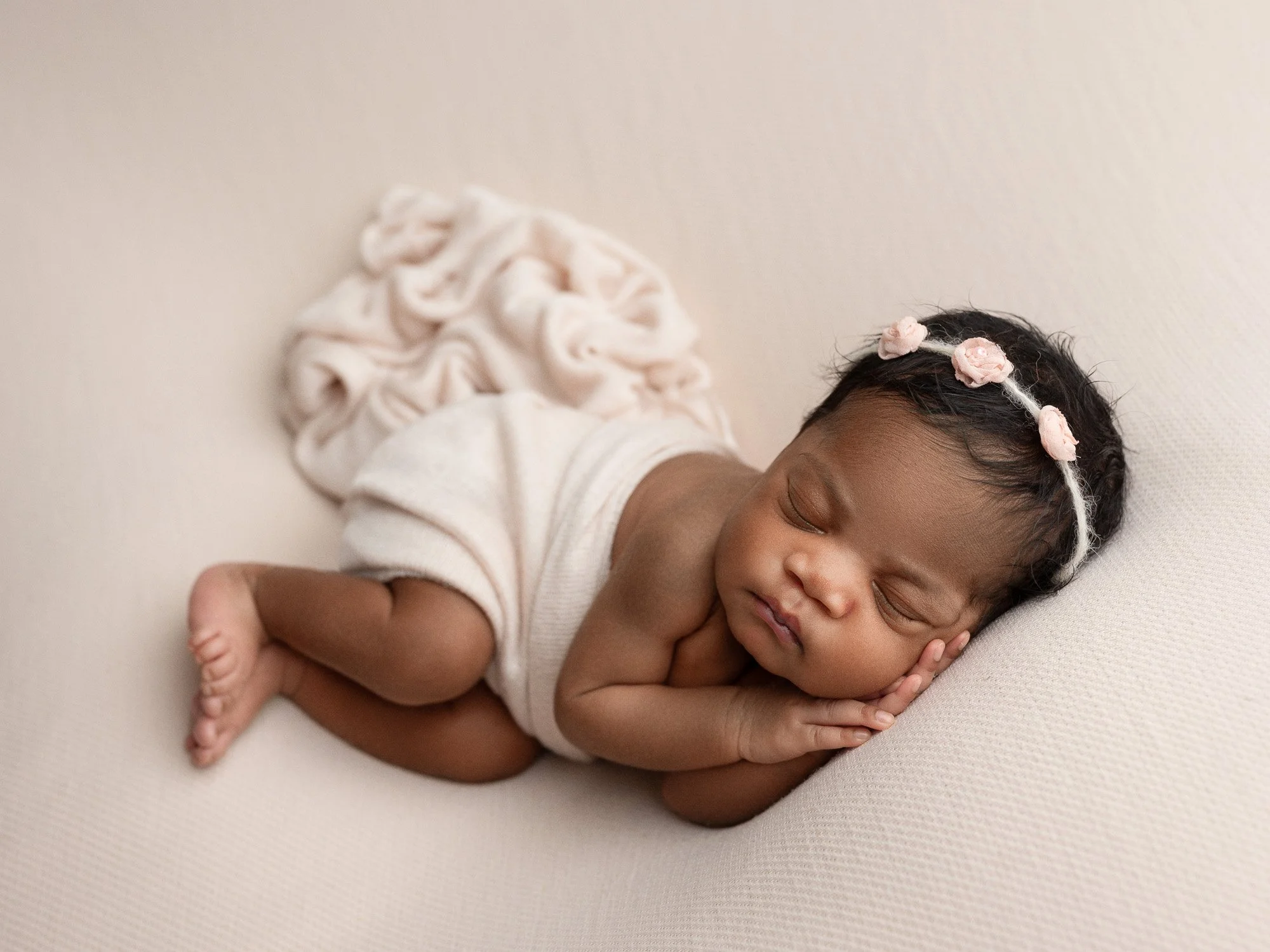 A sleeping newborn baby girl lying on her side on a soft, light-colored surface, wearing a headband with pink flowers, dressed in a light-colored outfit with a ruffled bottom.
