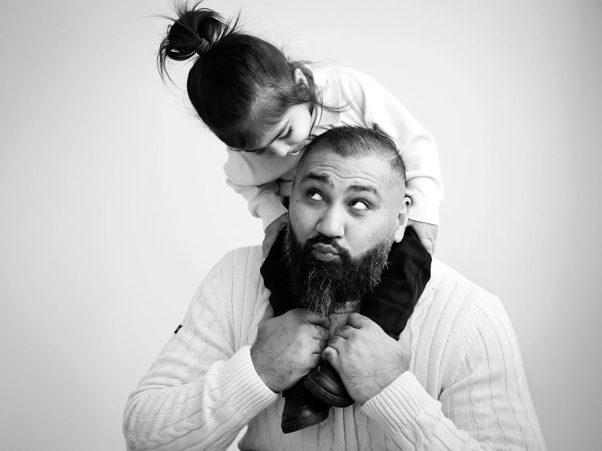 A man with a beard holding a young girl on his shoulders, both looking at each other playful and happy in a black-and-white photo.