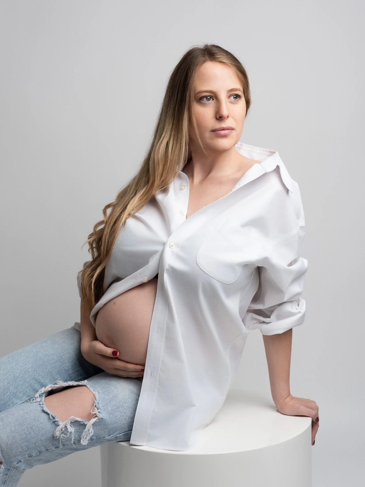 A pregnant woman with long wavy blonde hair, wearing a white button-up shirt and ripped jeans, sitting on a white cylindrical surface against a plain gray background.
