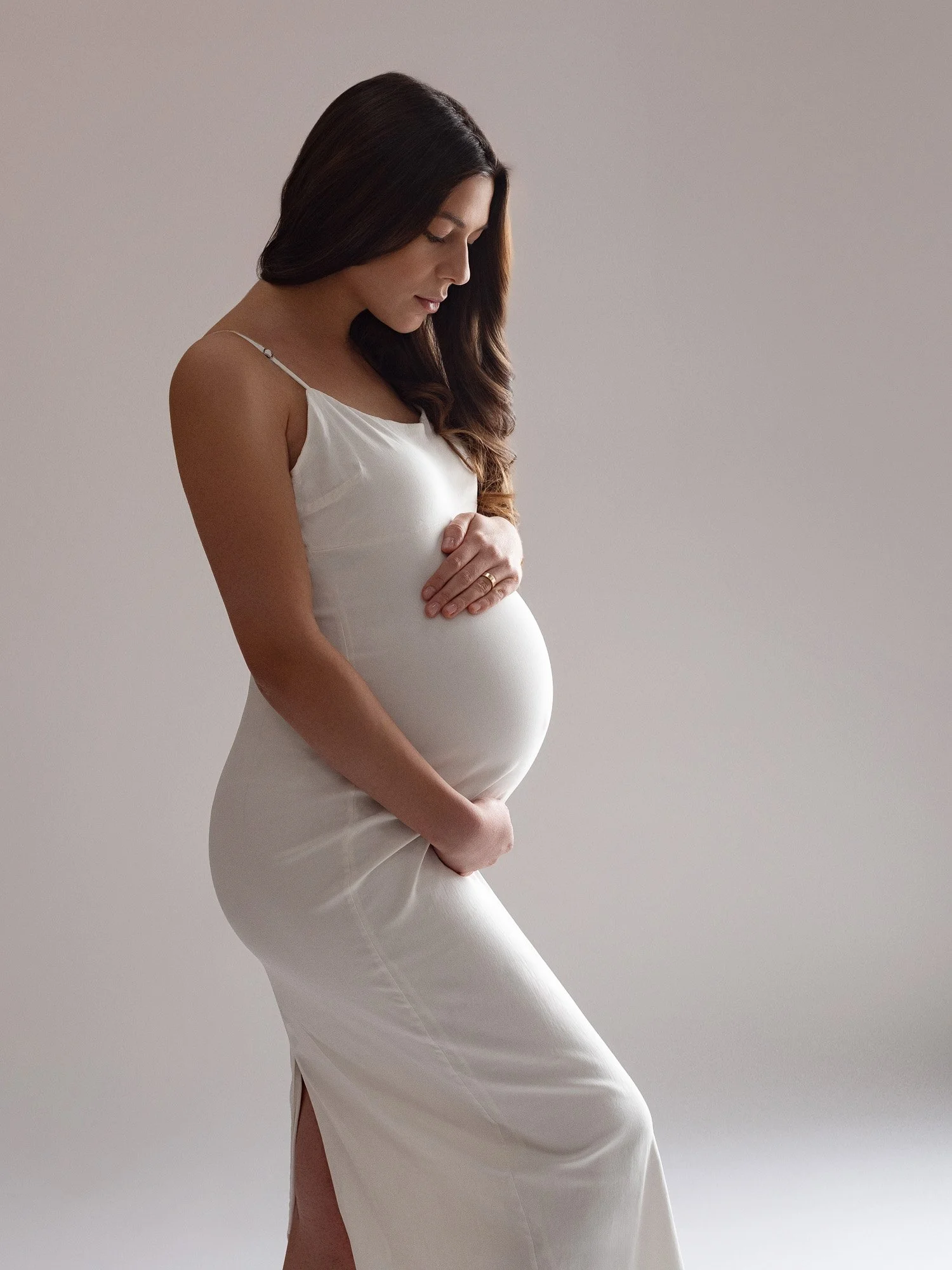 A pregnant woman with long brown hair wearing a white dress, gently holding her belly with both hands, looking down, standing against a plain light gray background.