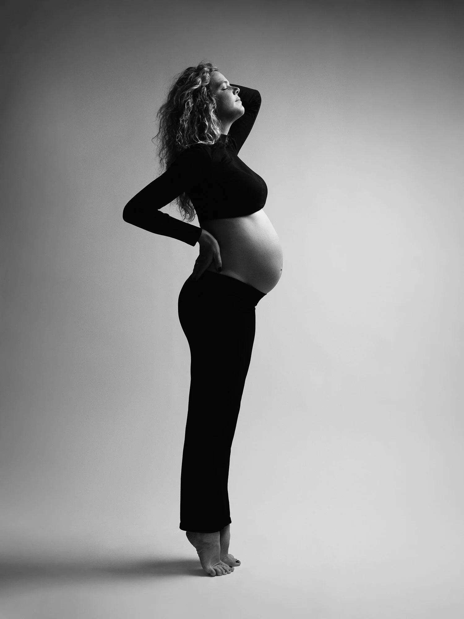 A pregnant woman with curly hair standing in profile with her hand on her hip and her eyes closed.