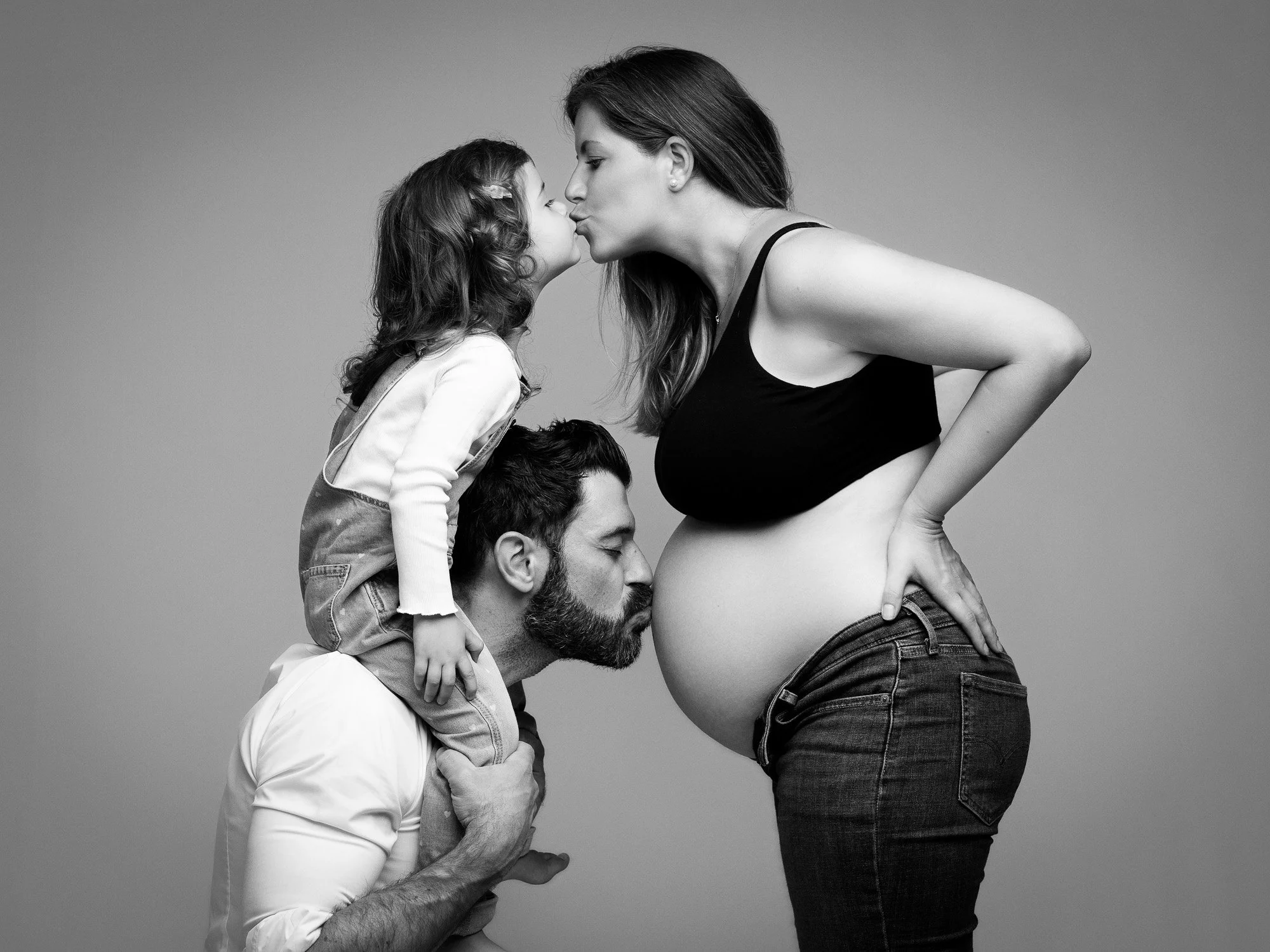A pregnant woman with long hair and a black top kissing a young girl, who is standing on a man's shoulders, on the forehead. The man has a beard and is holding the girl's legs. The scene is in black and white.