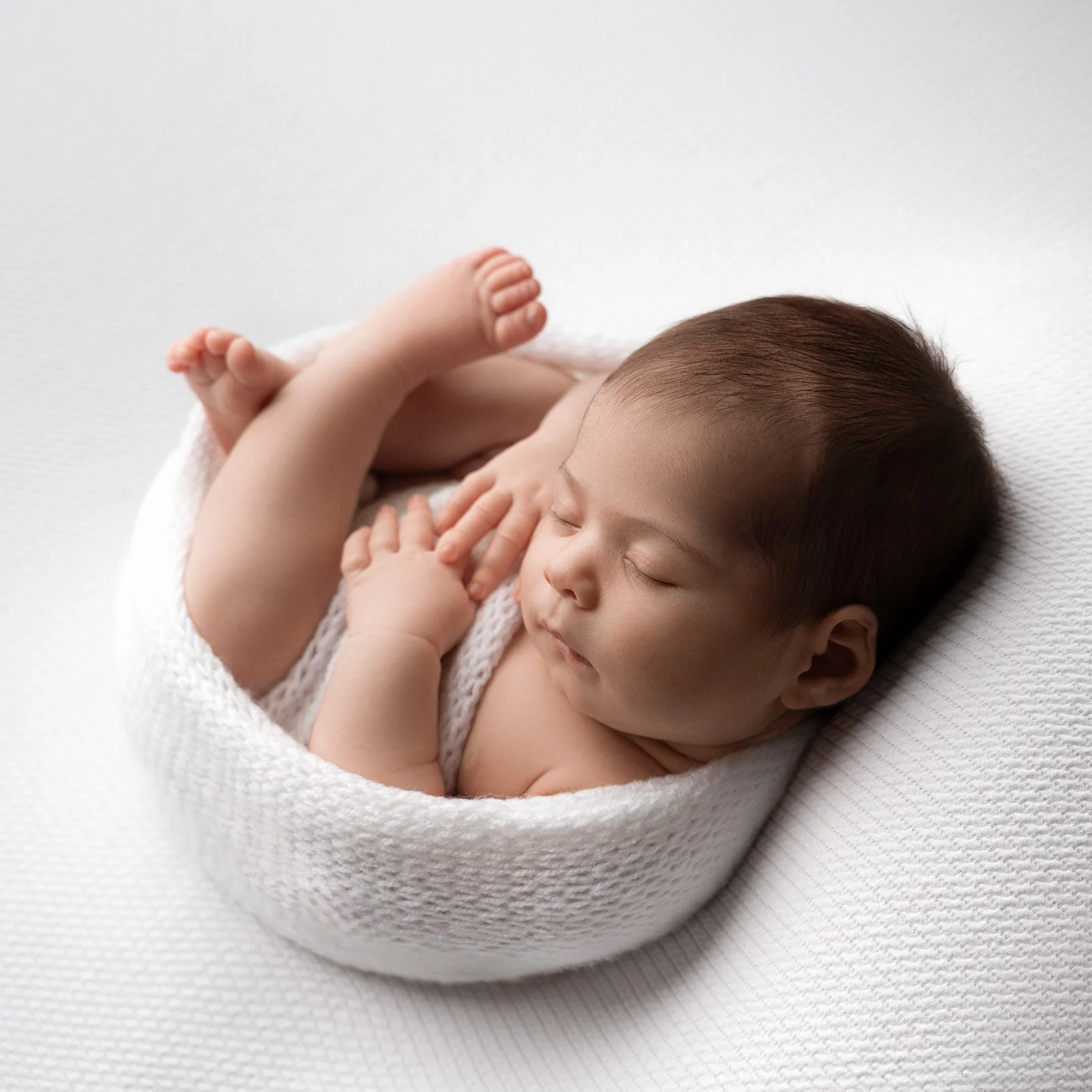 A sleeping newborn baby lying in a white knitted cocoon on a white textured surface.