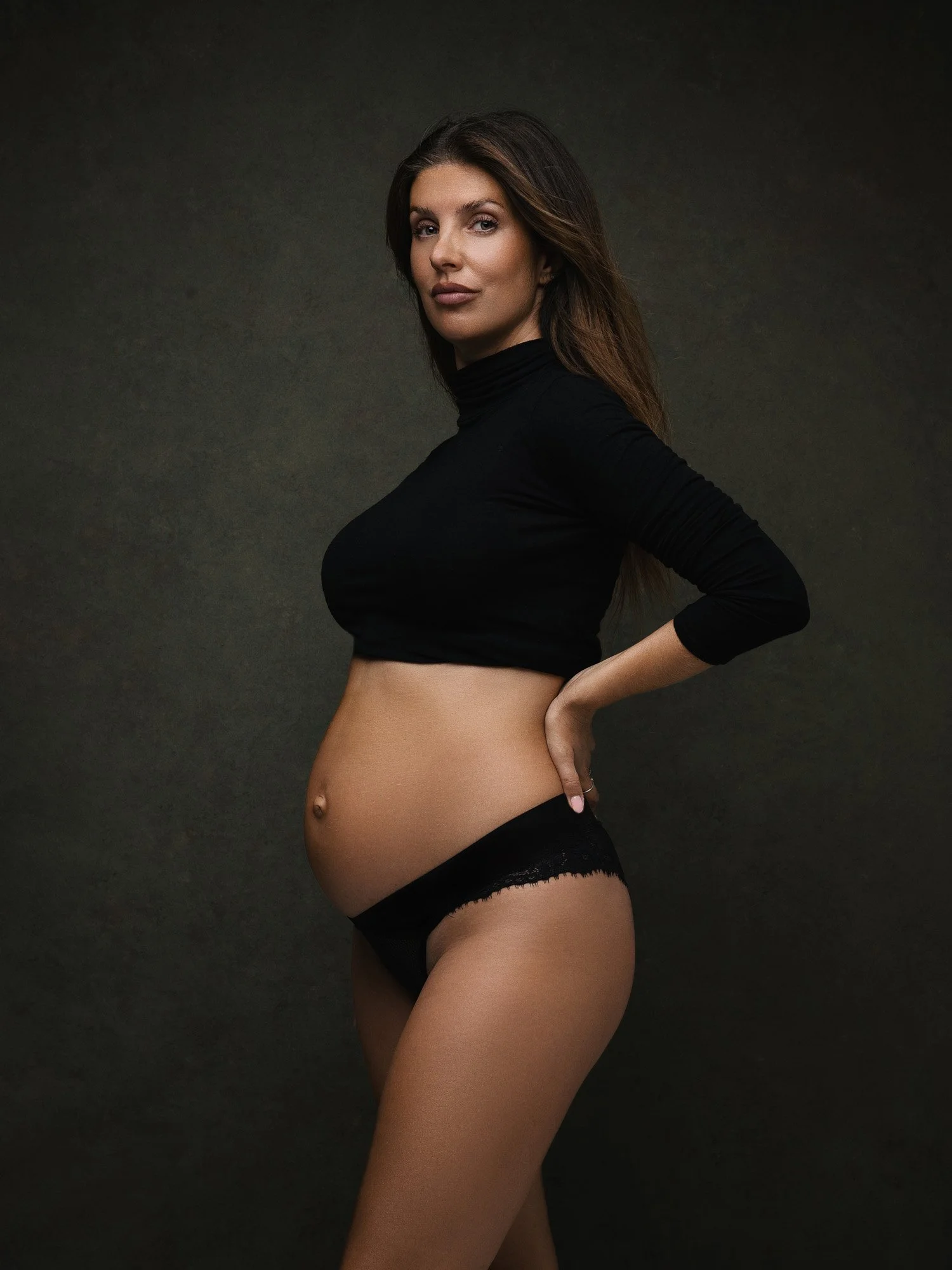A pregnant woman with long brown hair wearing a black turtleneck and black lace underwear, standing against a dark background.