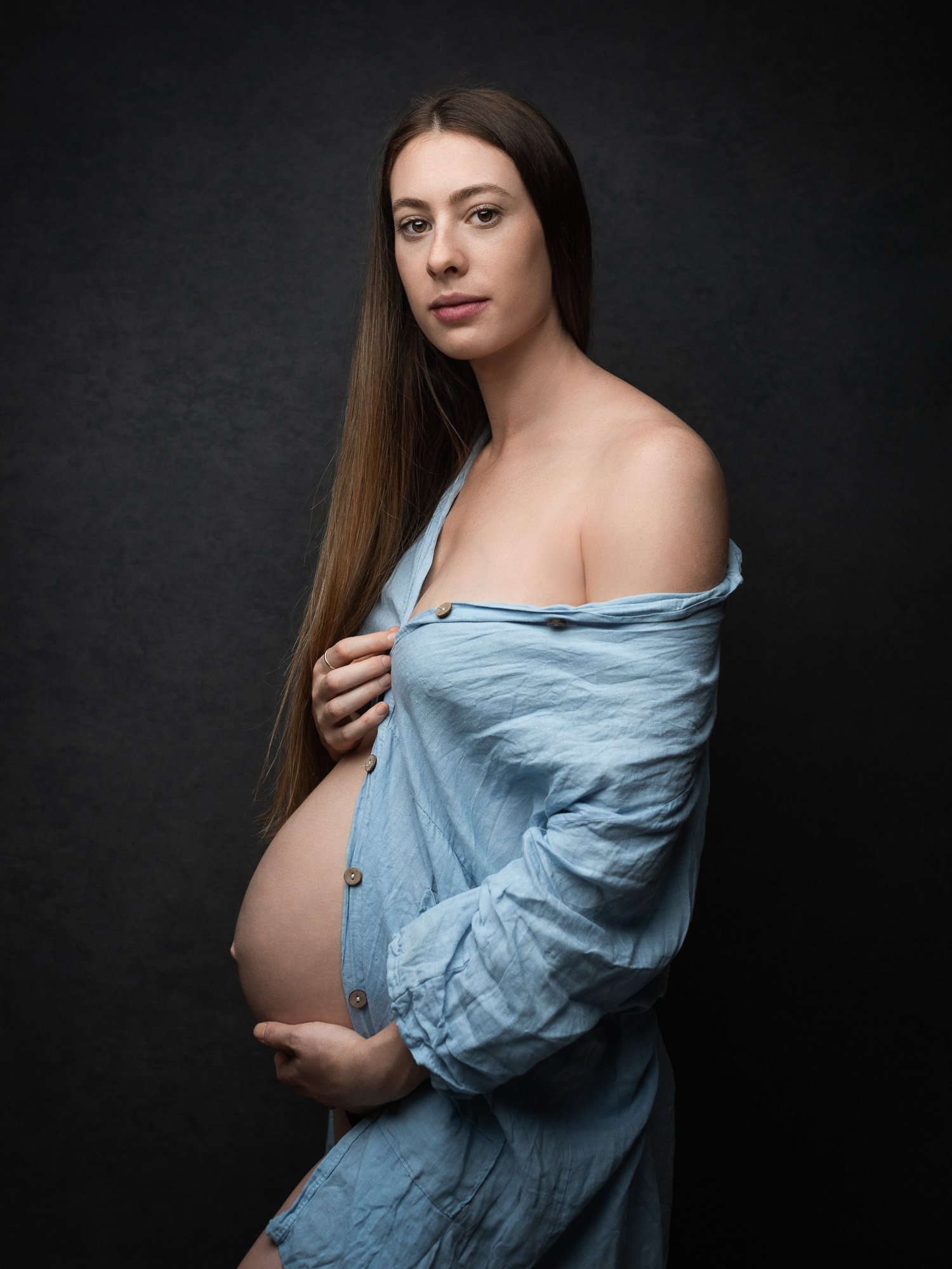 A pregnant woman with long brown hair, wearing a partially unbuttoned blue shirt, stands against a dark background, gazing at the camera.