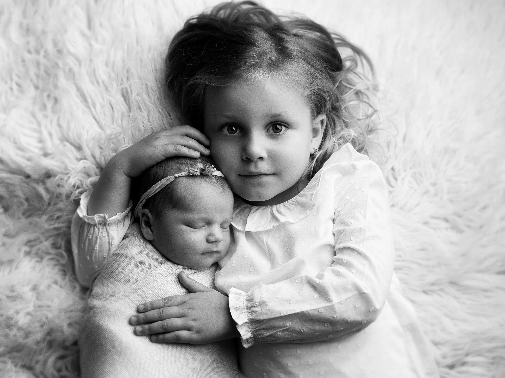 A young girl with curly hair holding a sleeping newborn girl on a fluffy surface.