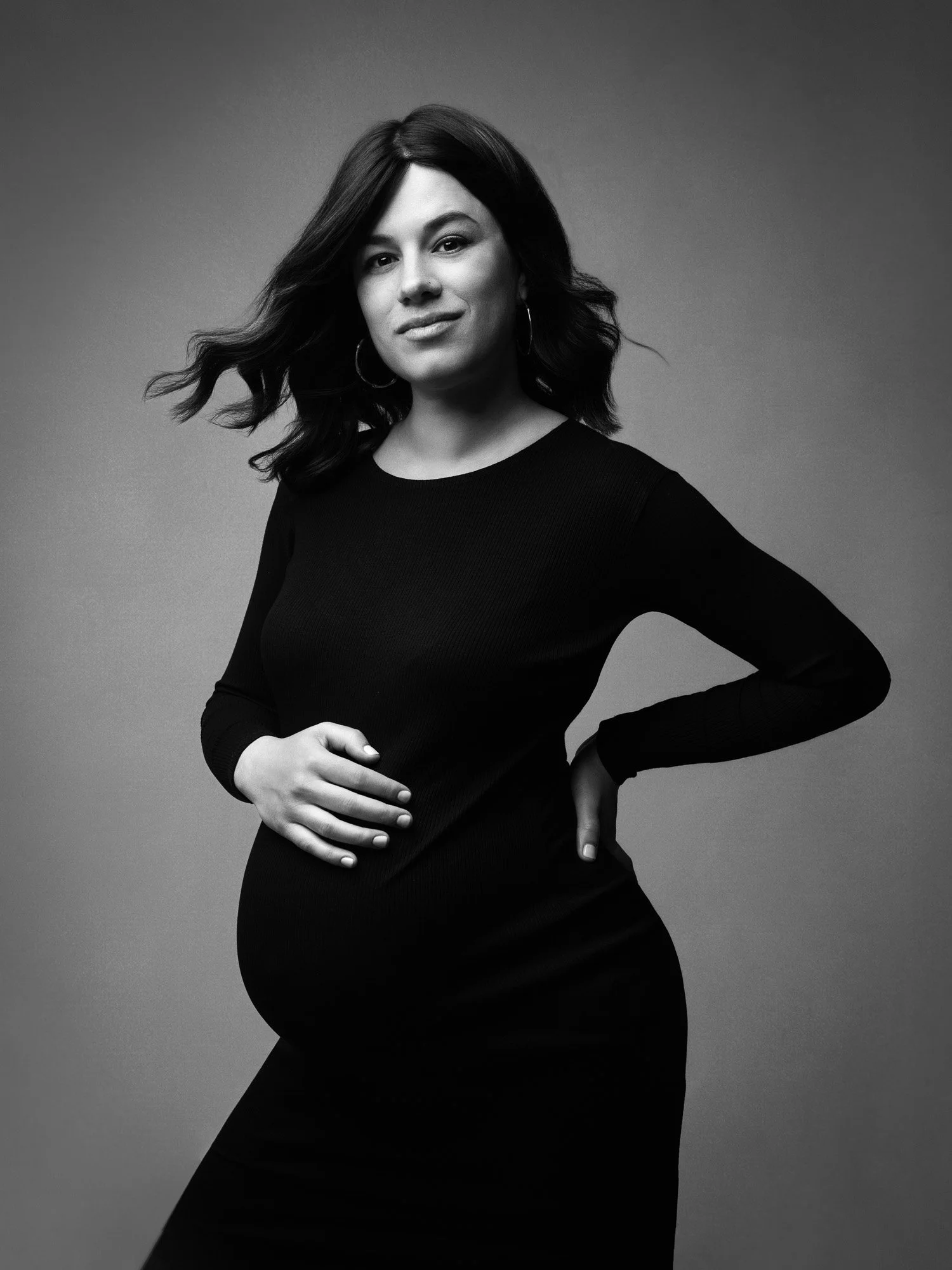 Black and white portrait of a woman with dark, wavy hair wearing hoop earrings and a long-sleeved black dress, standing with one hand on her hip and the other gently resting on her stomach.