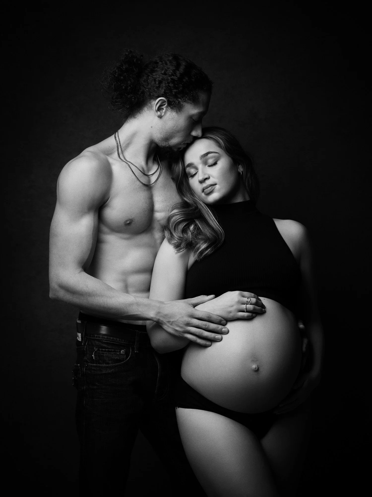 Black and white photo of a pregnant woman with long hair, clothed in a sleeveless top and underwear, being embraced by a shirtless man with long, curly hair, both with eyes closed, against a dark background.