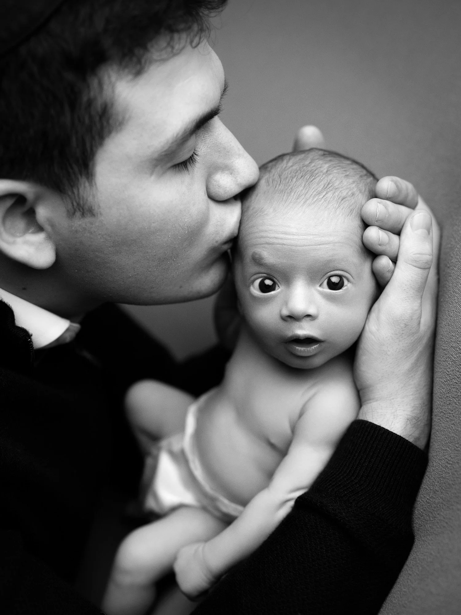 A man is kissing a baby on the forehead, holding the baby gently with one hand, while the baby's wide eyes look directly at the camera. The image is in black and white.