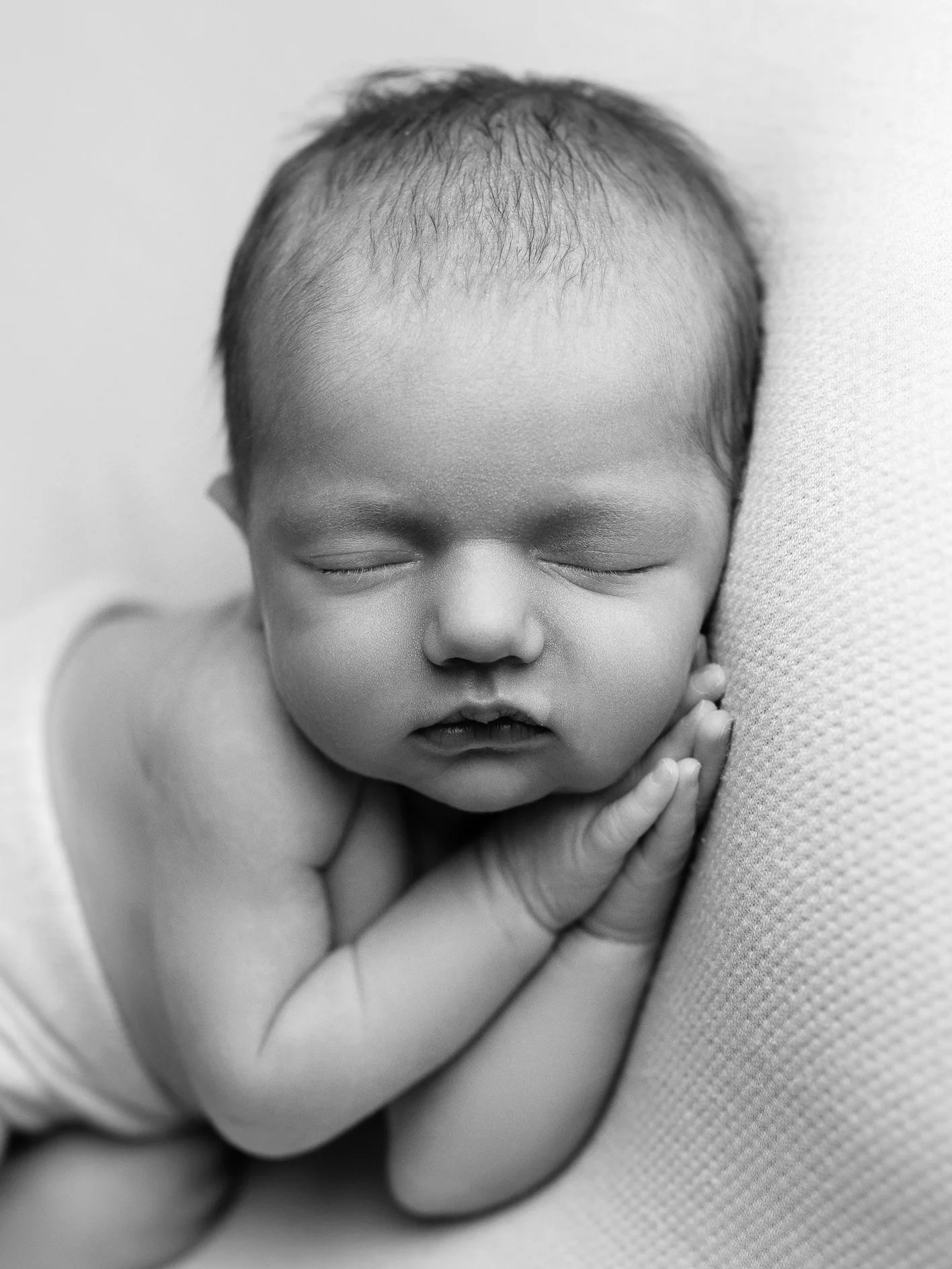 Close-up black and white photo of a sleeping baby with hands near face, resting on a textured surface.