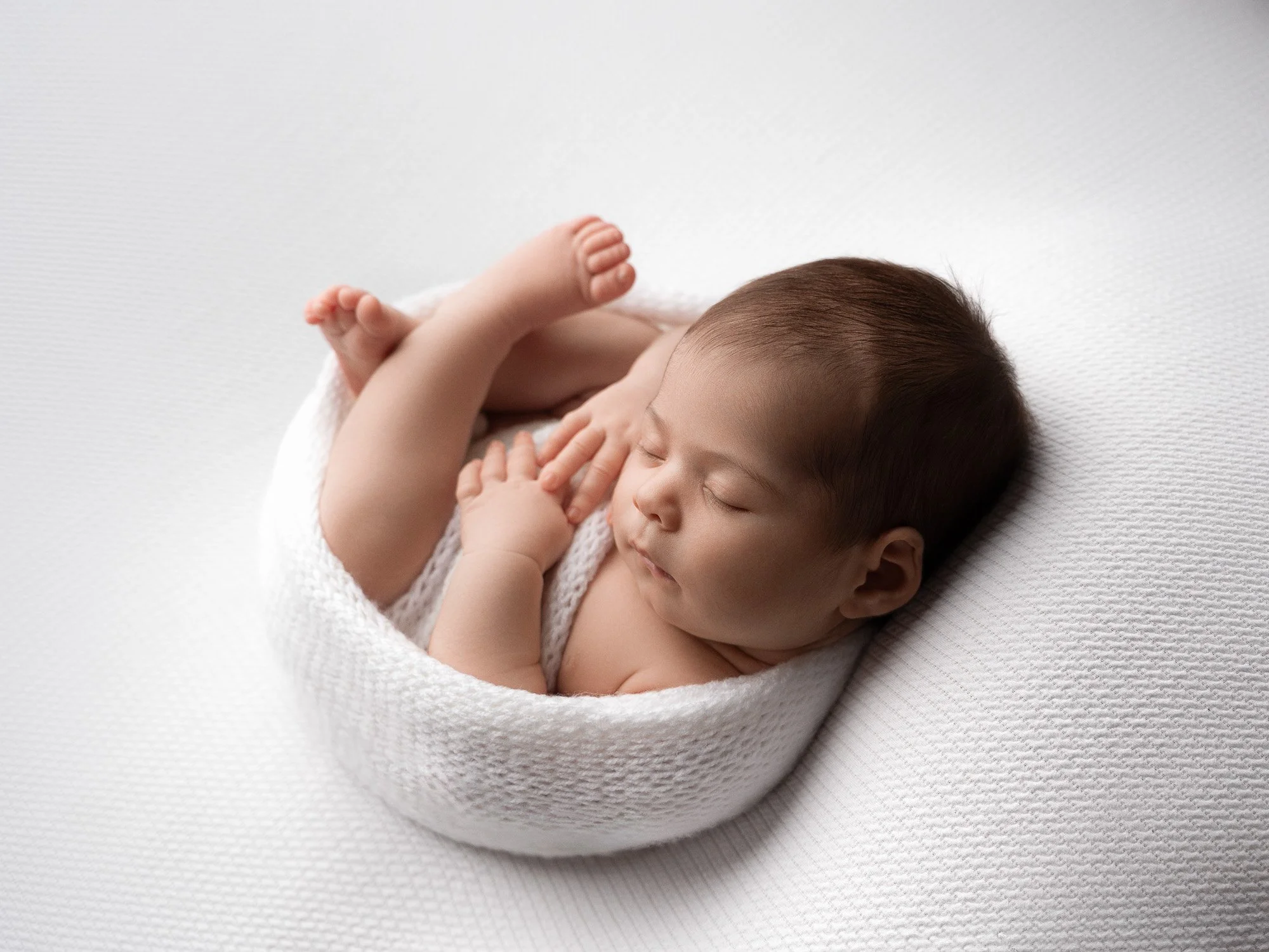 A sleeping baby curled up on a white textured blanket, wrapped in a white knitted blanket.