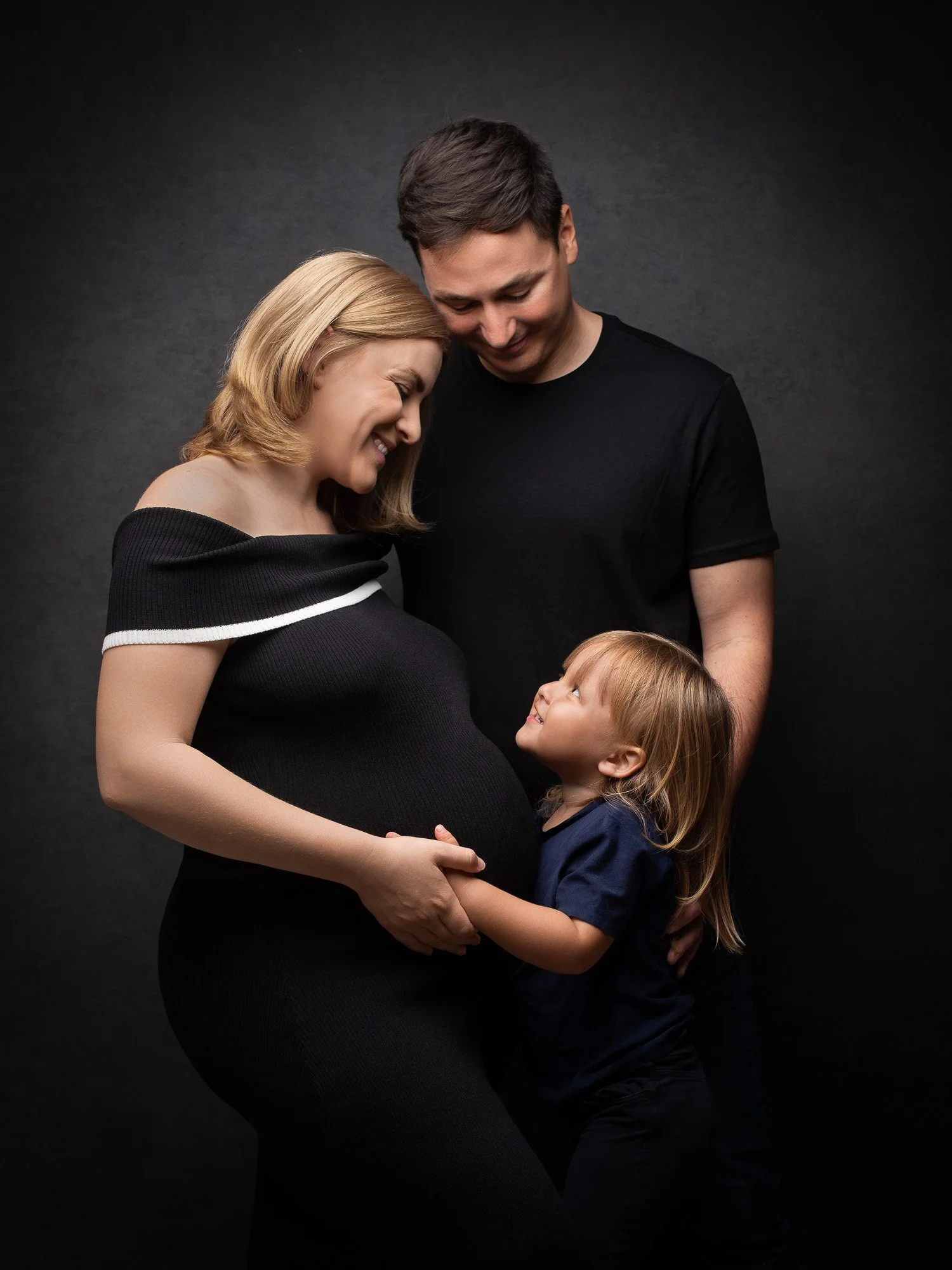 A pregnant woman with blonde hair in black dress with white stripe, a man with dark hair in a black shirt, and a young girl with red hair in a blue shirt are smiling and touching each other's hands against a dark background.