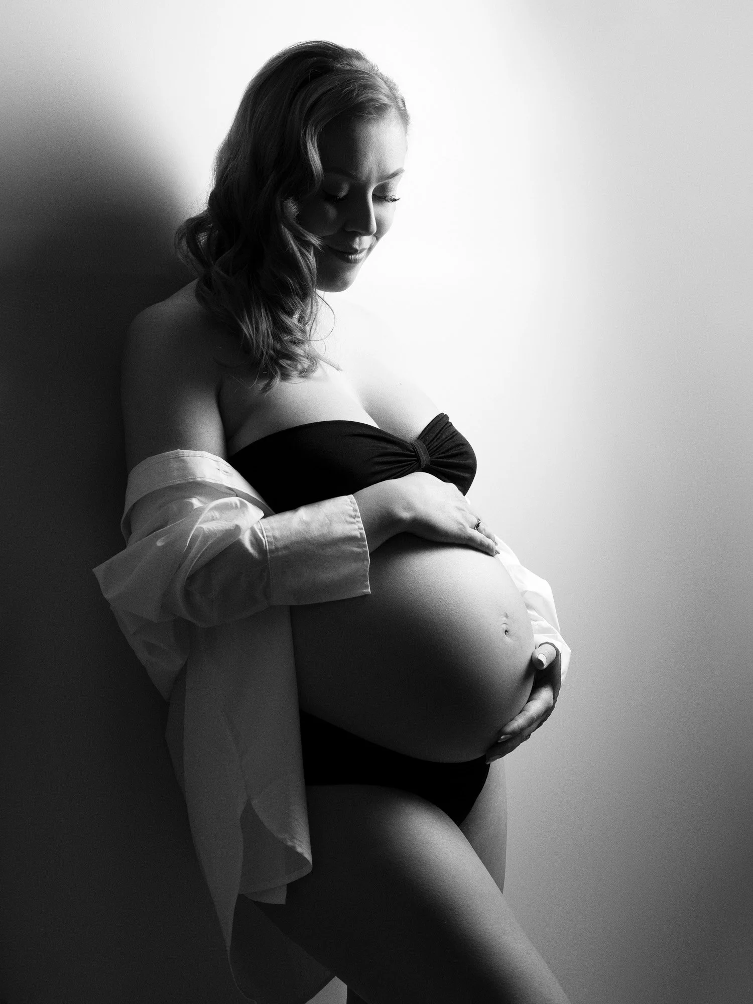 Black and white photo of a pregnant woman smiling, wearing a black bandeau top and an unbuttoned shirt, holding her belly with one hand and a small object in the other.