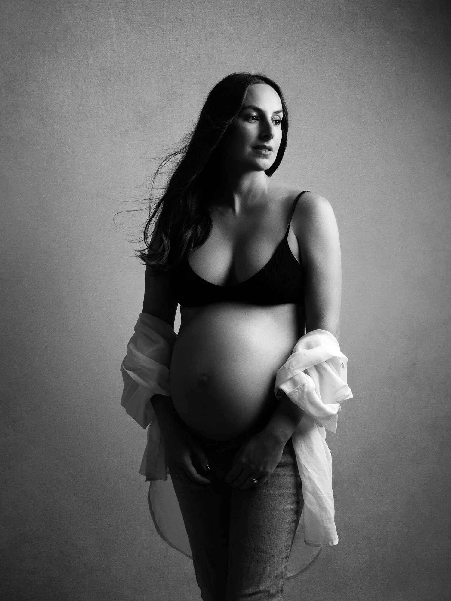Black and white portrait of a pregnant woman with long hair, wearing a bra and jeans, with a shirt draped over her shoulders, standing against a plain background.