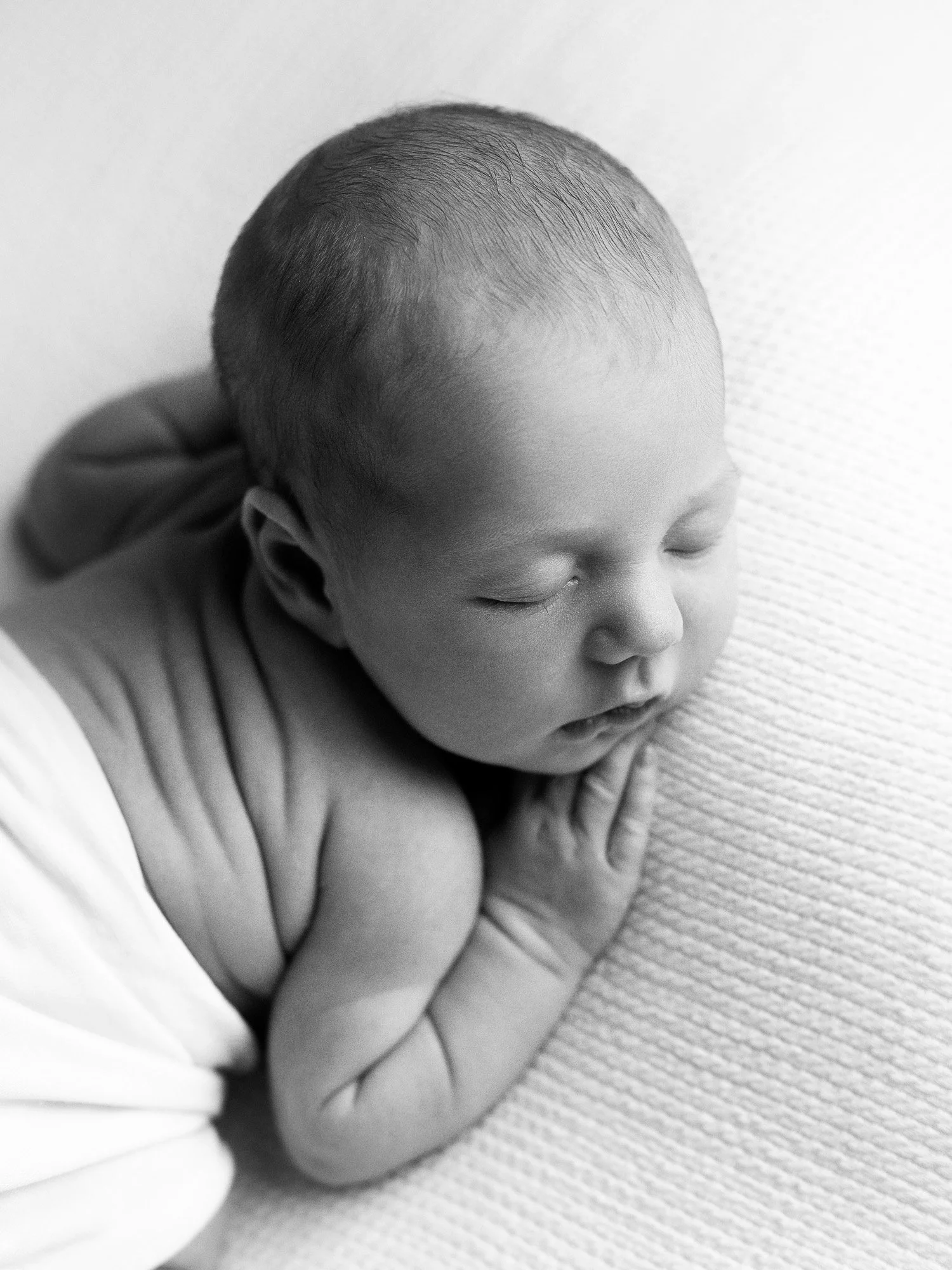 Black and white photo of a sleeping baby lying on their side, with their hands tucked under their head, on a textured surface.