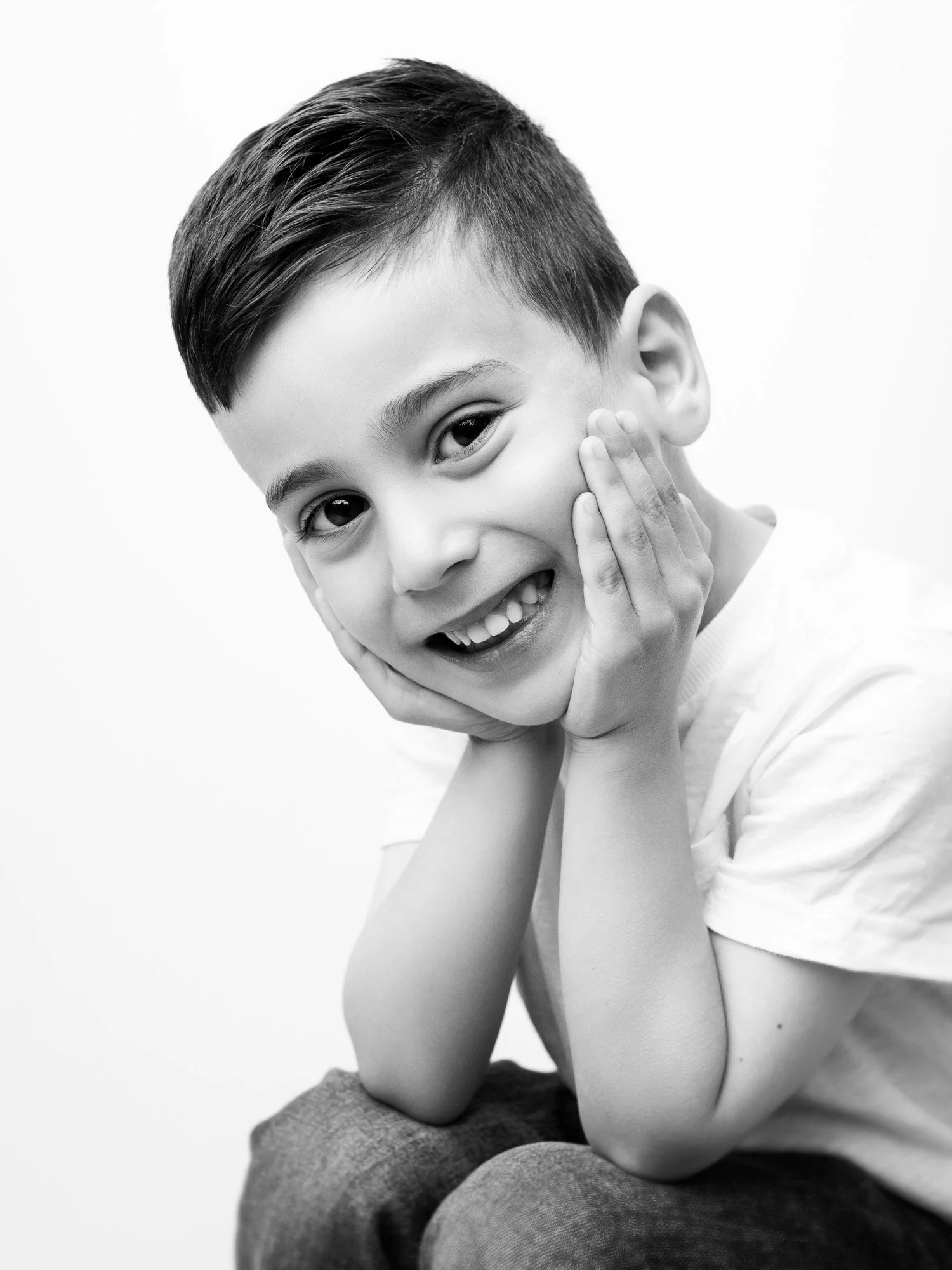 A smiling young boy with short hair, holding his face with both hands, in a black and white portrait.