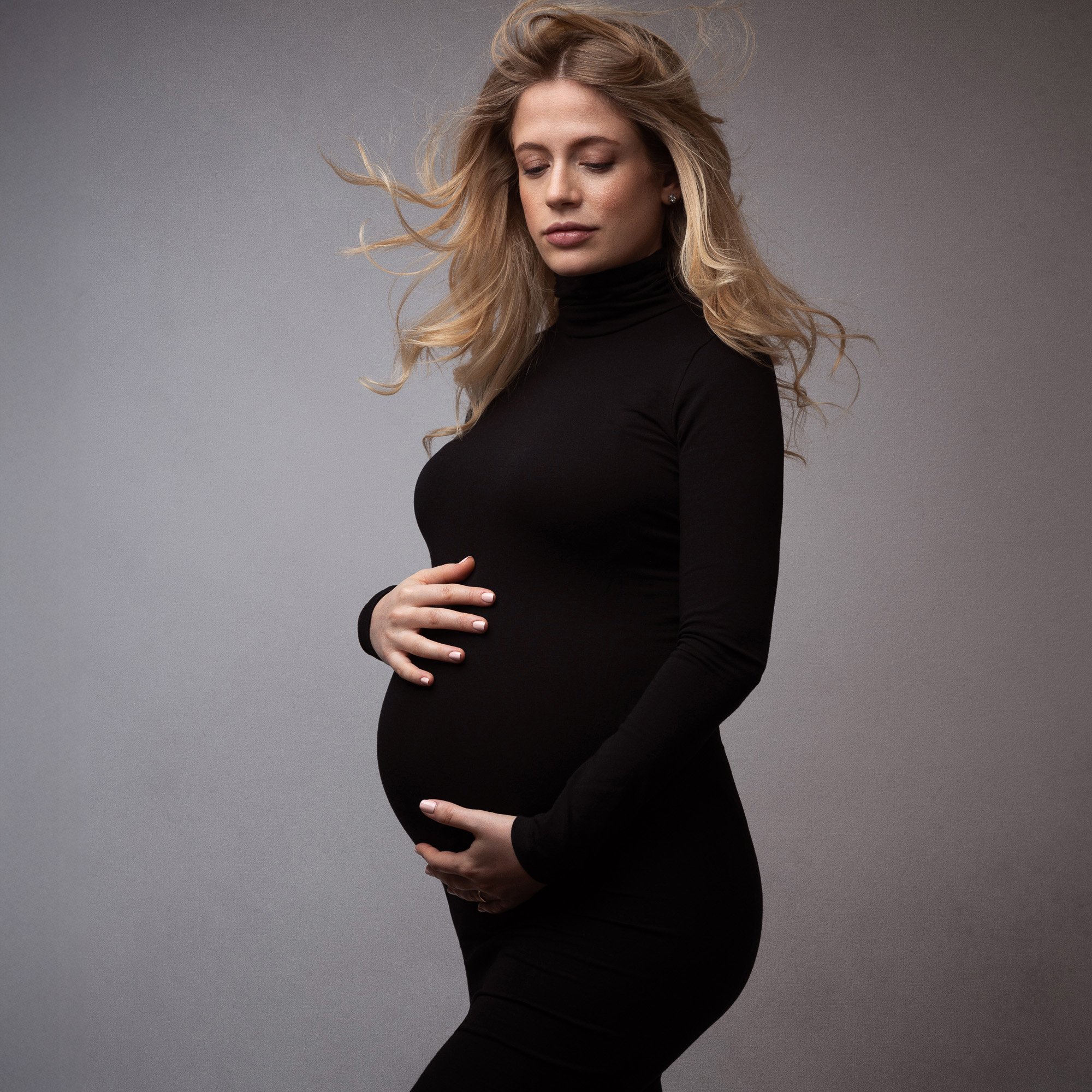 A pregnant woman with long blonde hair, wearing a black turtleneck dress, stands against a plain gray background, gently holding her belly with both hands and looking down with a serene expression.