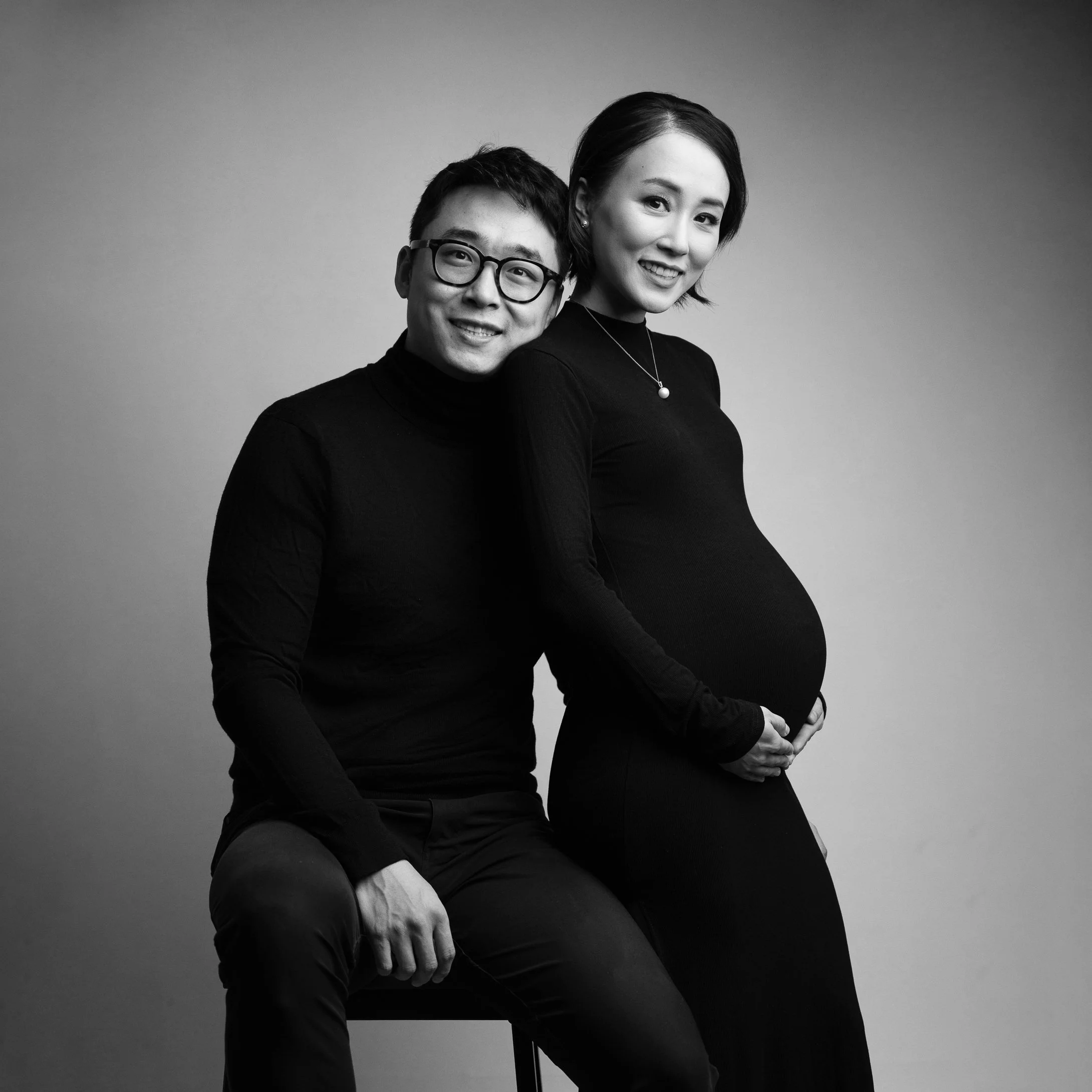 Black and white portrait of a couple, with the woman pregnant, both smiling at the camera, against a plain background.