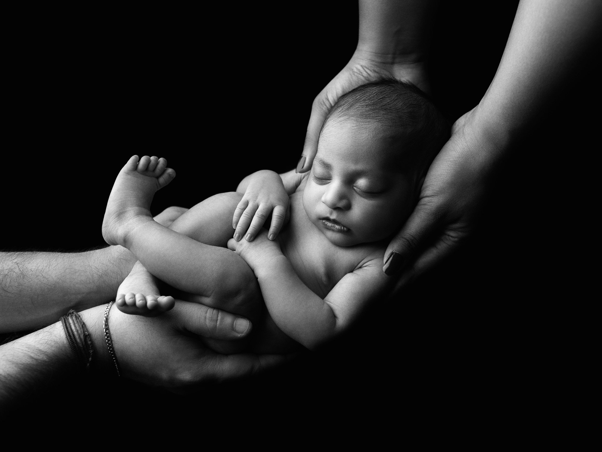 Black and white photo of a newborn baby being gently held in multiple hands, with one hand supporting the head and another cradling the body, against a dark background.