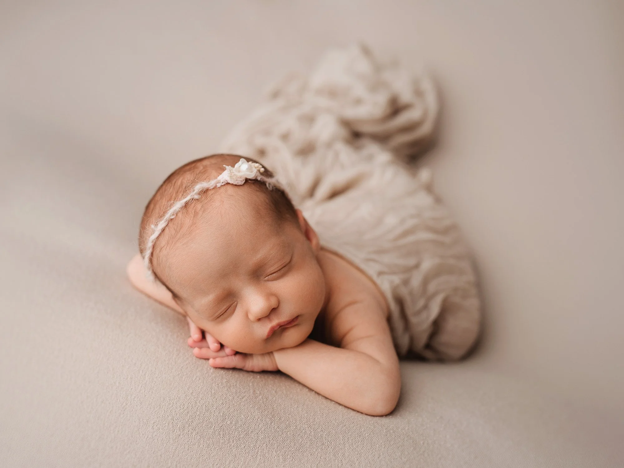 A sleeping newborn baby with a delicate headband, lying on a beige surface, wrapped in a soft beige blanket.