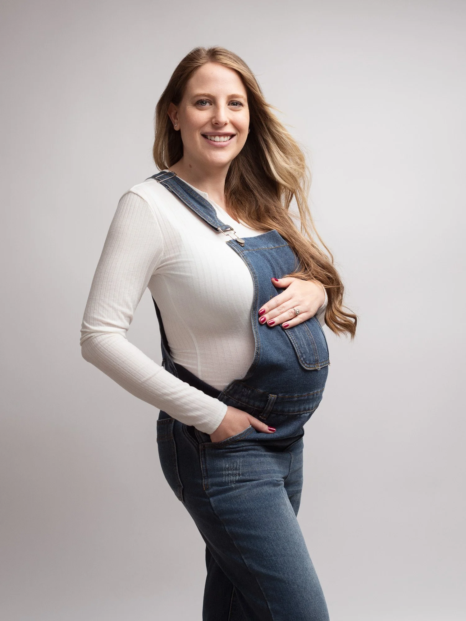Pregnant woman with long hair smiling, wearing a white long-sleeve shirt and blue denim overalls, standing against a plain light-colored background.