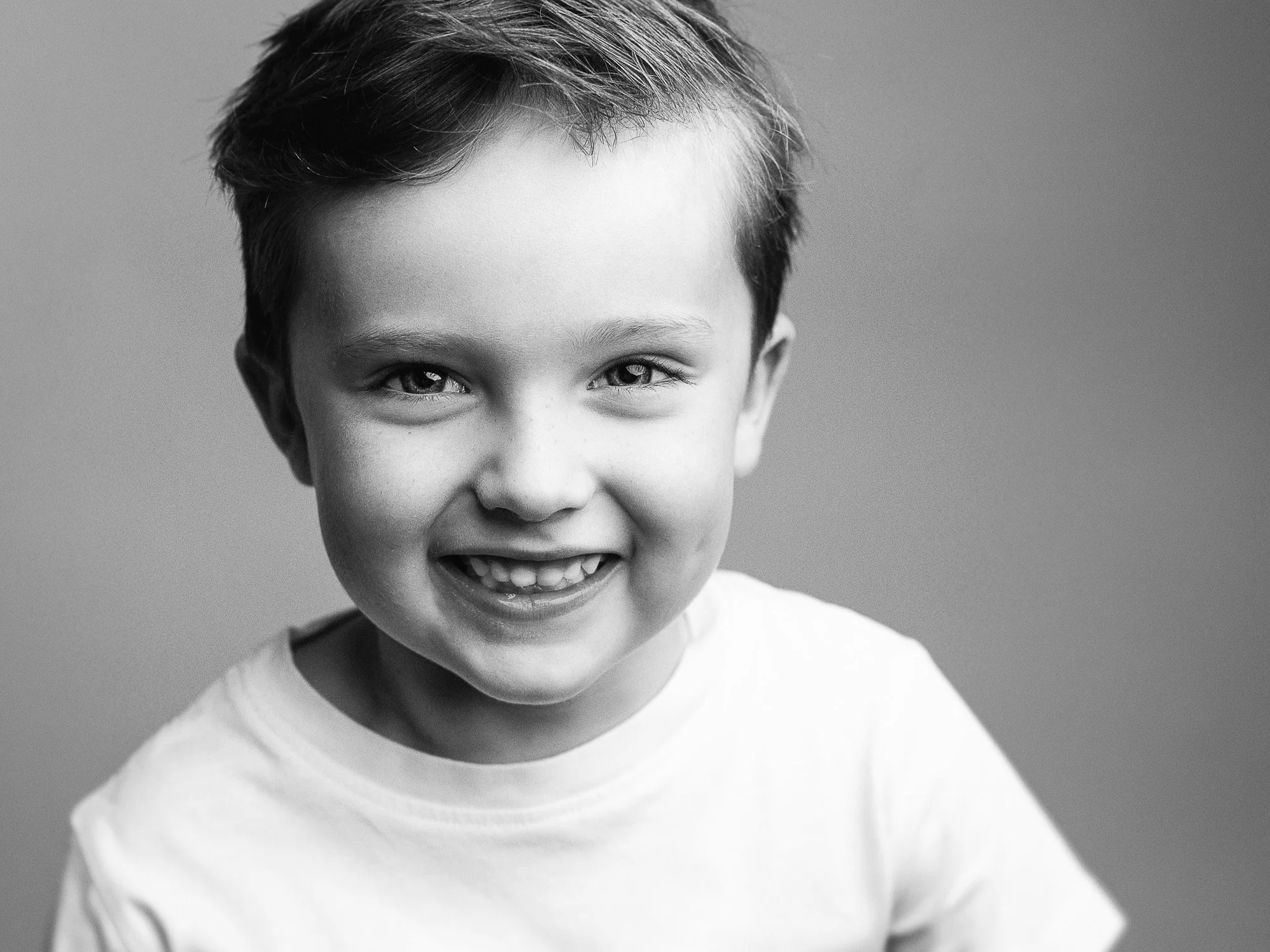 A smiling young boy with short hair, wearing a white T-shirt, in a black and white portrait.