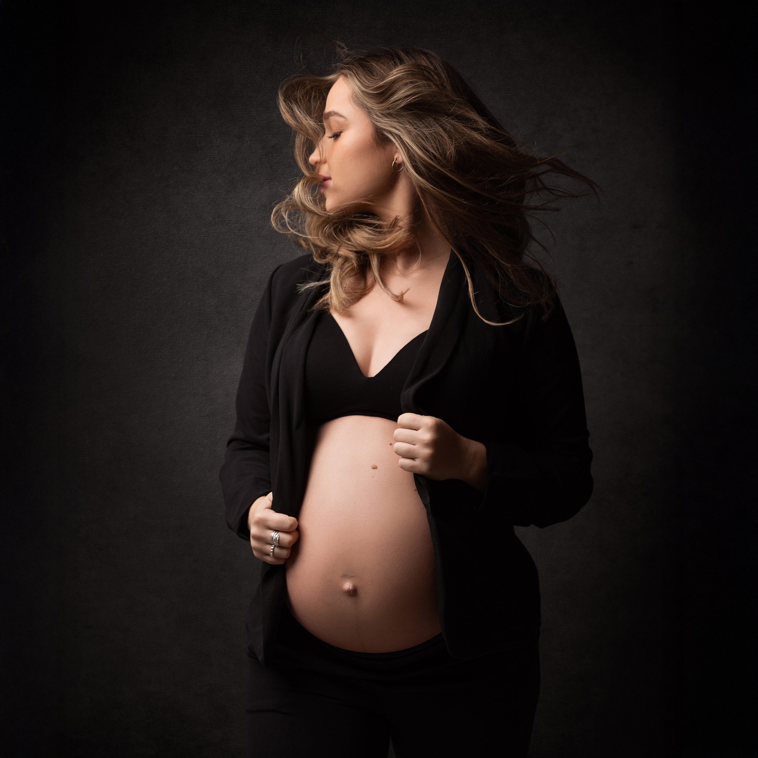 A pregnant woman with wavy hair wearing a black bra and open black jacket, posing against a dark background.