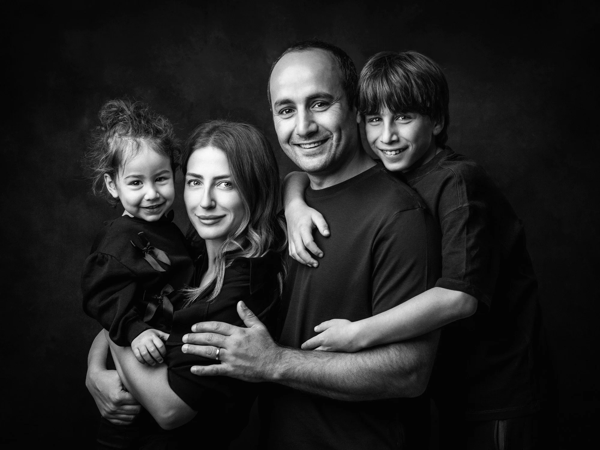 Black and white portrait of a smiling family of four: a father, a mother, a daughter, and a son, all dressed in dark clothing, with the children on the parents' laps, against a dark background.