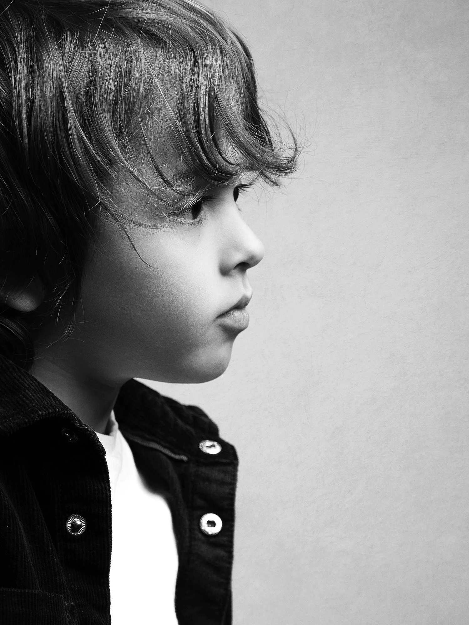 A black and white side profile of a young boy with wavy hair, wearing a dark jacket with metal buttons.