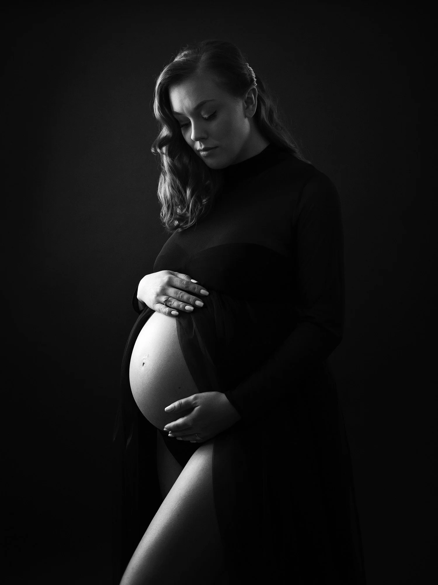 A pregnant woman in a black dress gently holding her belly against a dark background.