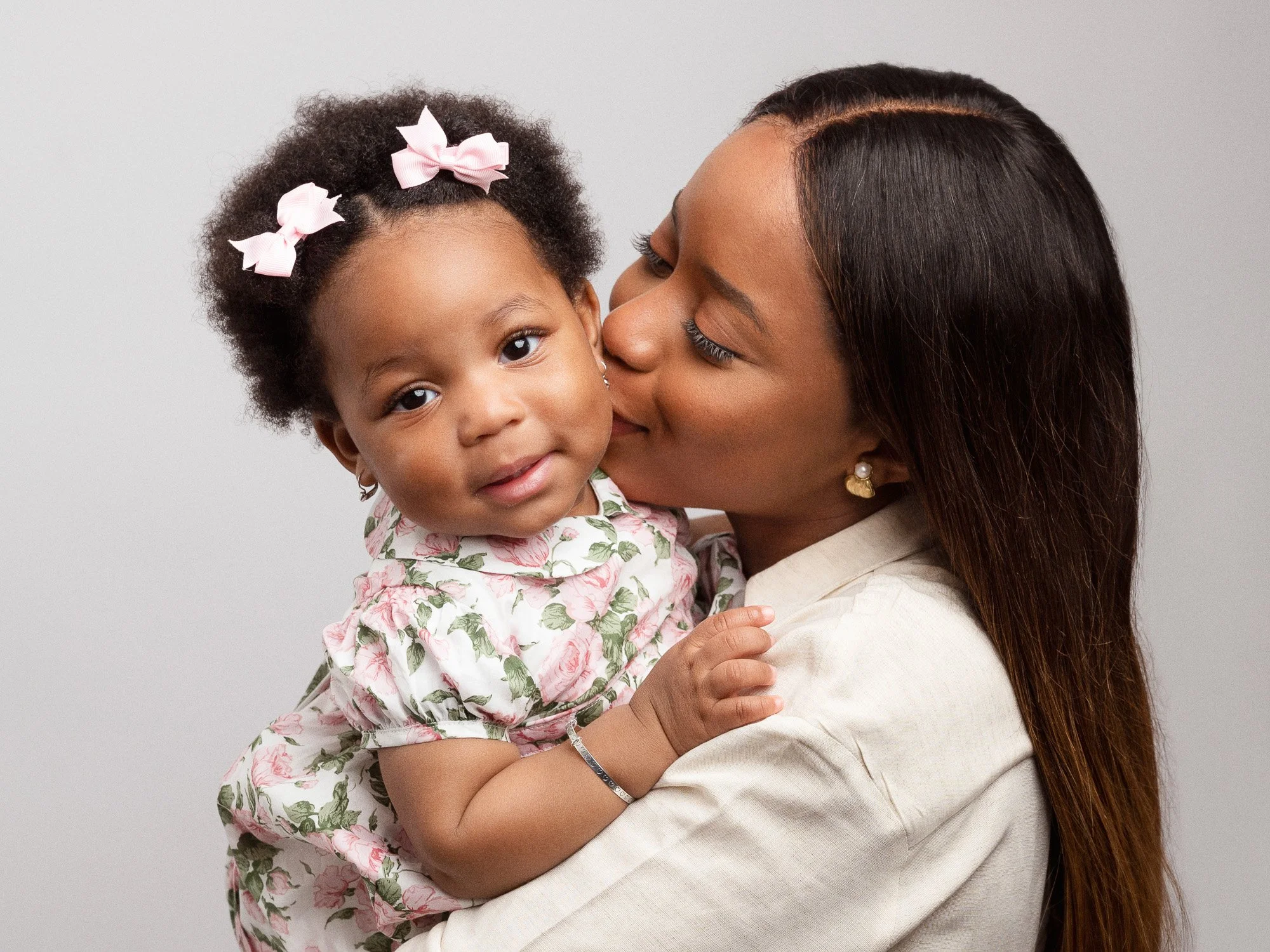 A woman with long dark hair and pearl earrings holding a young girl with curly hair and pink bows, both smiling, against a plain gray background.