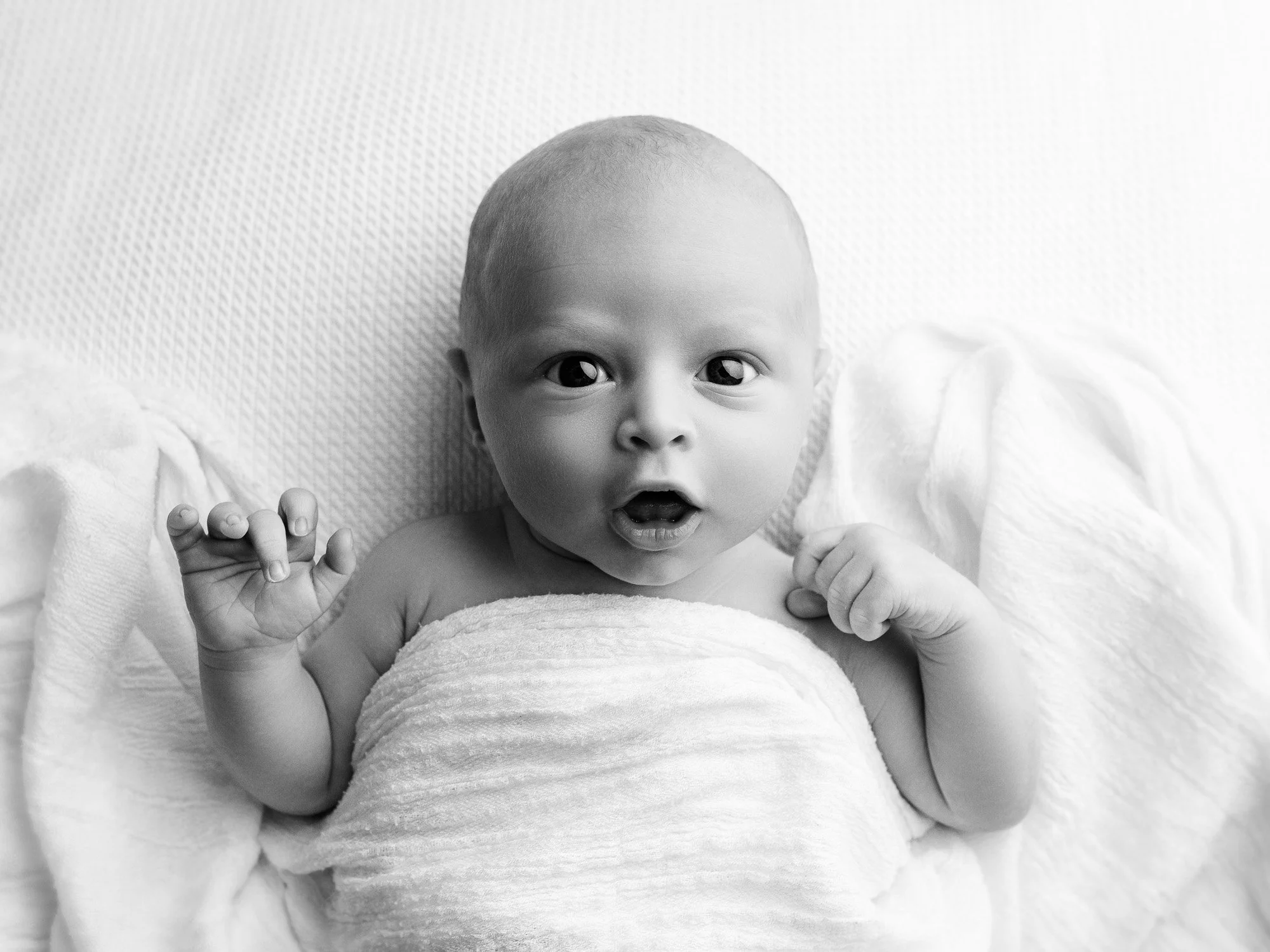 A black and white photo of a baby lying on a soft surface, wrapped in a towel, with wide eyes and an open mouth, looking surprised or curious.