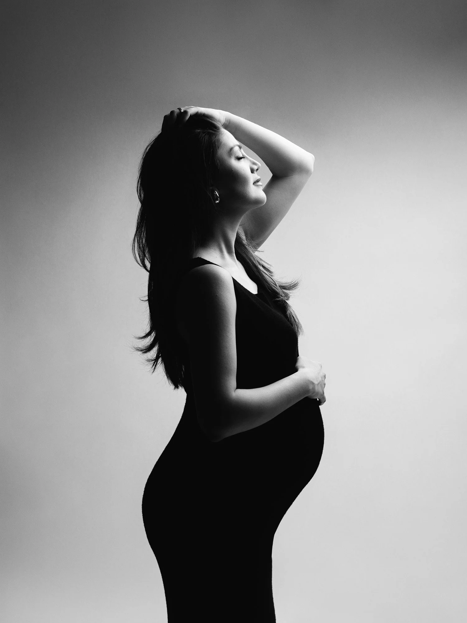 A pregnant woman in a black dress smiling with her hand on her head in a black and white photo.