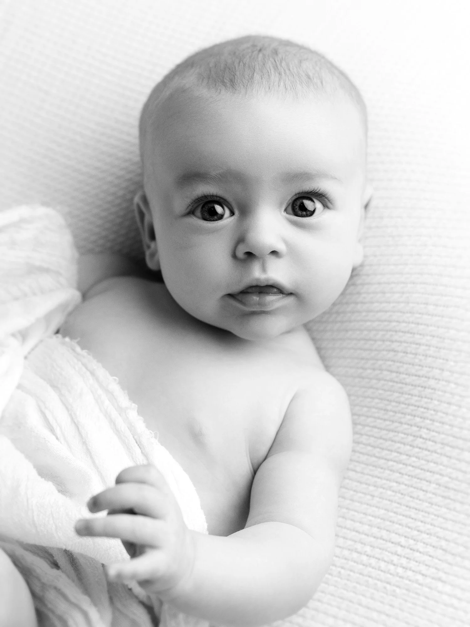 Black and white photo of a baby with big eyes looking at the camera, held in a towel, lying on a textured surface.