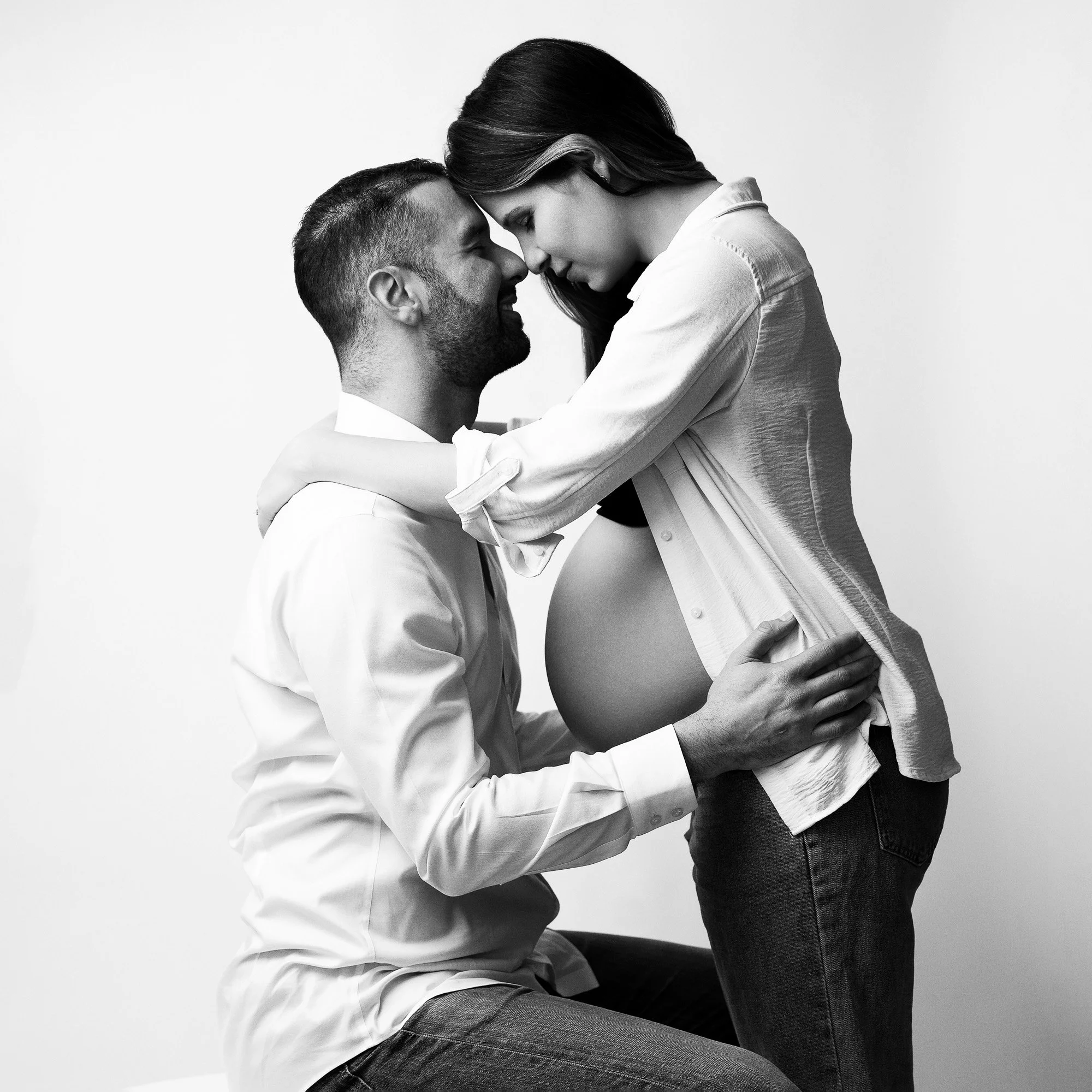 A black-and-white photo of a pregnant woman and a man, possibly the father. The man is kneeling, holding the woman's waist, and both are leaning their foreheads together, touching noses, with closed eyes. The woman is wearing a loose shirt that expos