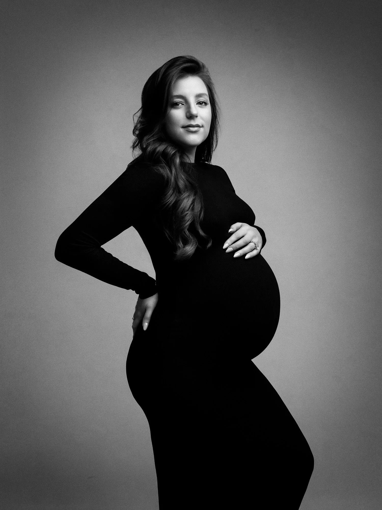 Black and white portrait of a pregnant woman with long wavy hair, wearing a form-fitting black dress, posing with her hand on her baby bump and looking confidently at the camera.